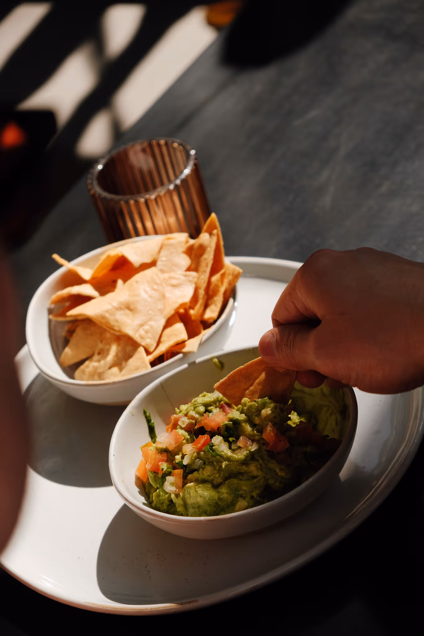 Hand dipping a tortilla chip into a bowl of guacamole with diced tomatoes and onions, next to a bowl of tortilla chips on a white plate.