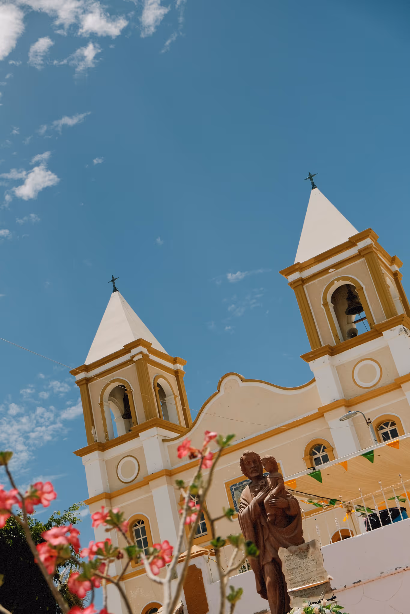 Yellow and white church with twin bell towers under a blue sky with scattered clouds and a statue of a saint holding a child in the foreground.