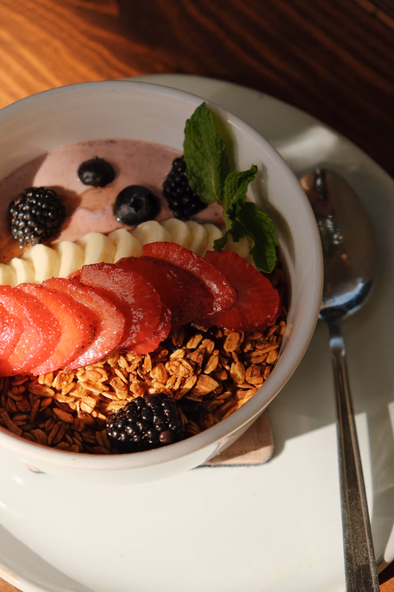 Bowl of granola topped with sliced strawberries, banana, blackberries, blueberries, and a sprig of mint on a white plate with a spoon.