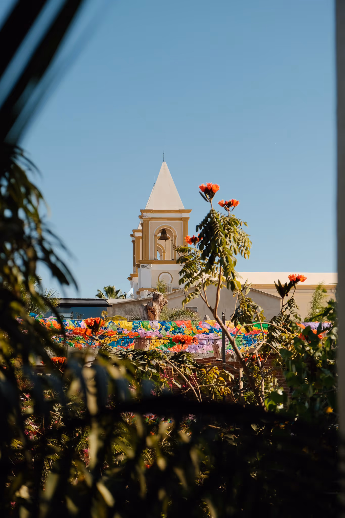 Bell tower of a beige church with colorful papel picado banners and green foliage in the foreground under a clear blue sky.