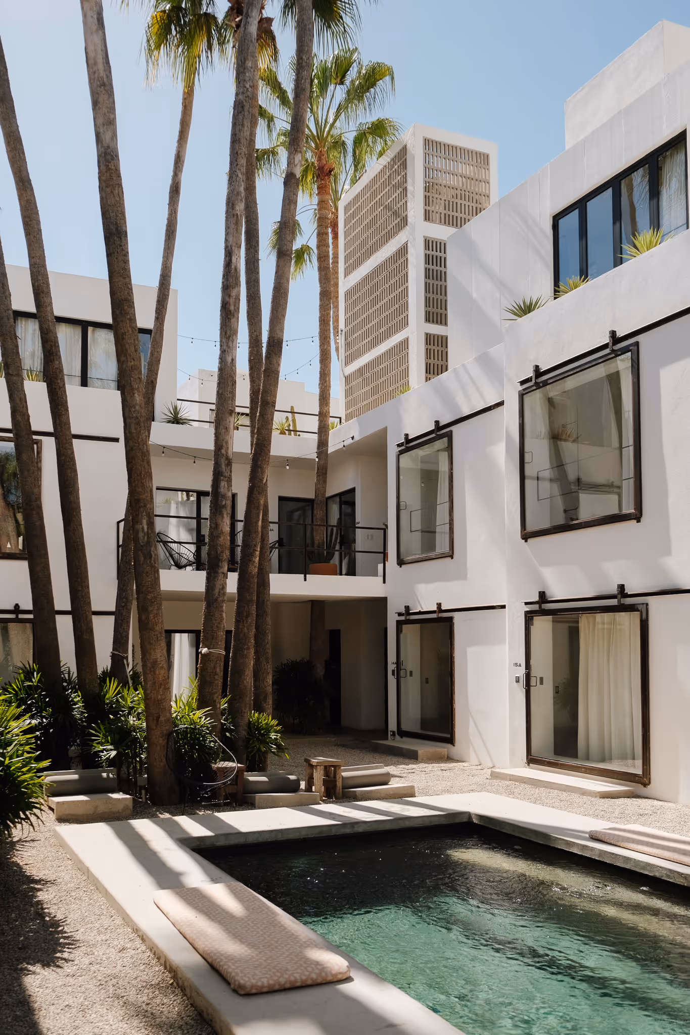 Modern white building surrounding a small rectangular pool with palm trees and lounge cushions under clear sky.
