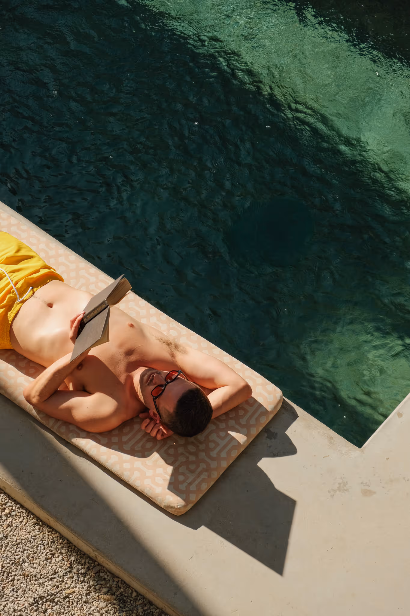 Man wearing yellow swim shorts lounging on a patterned poolside cushion reading a book next to deep green water.