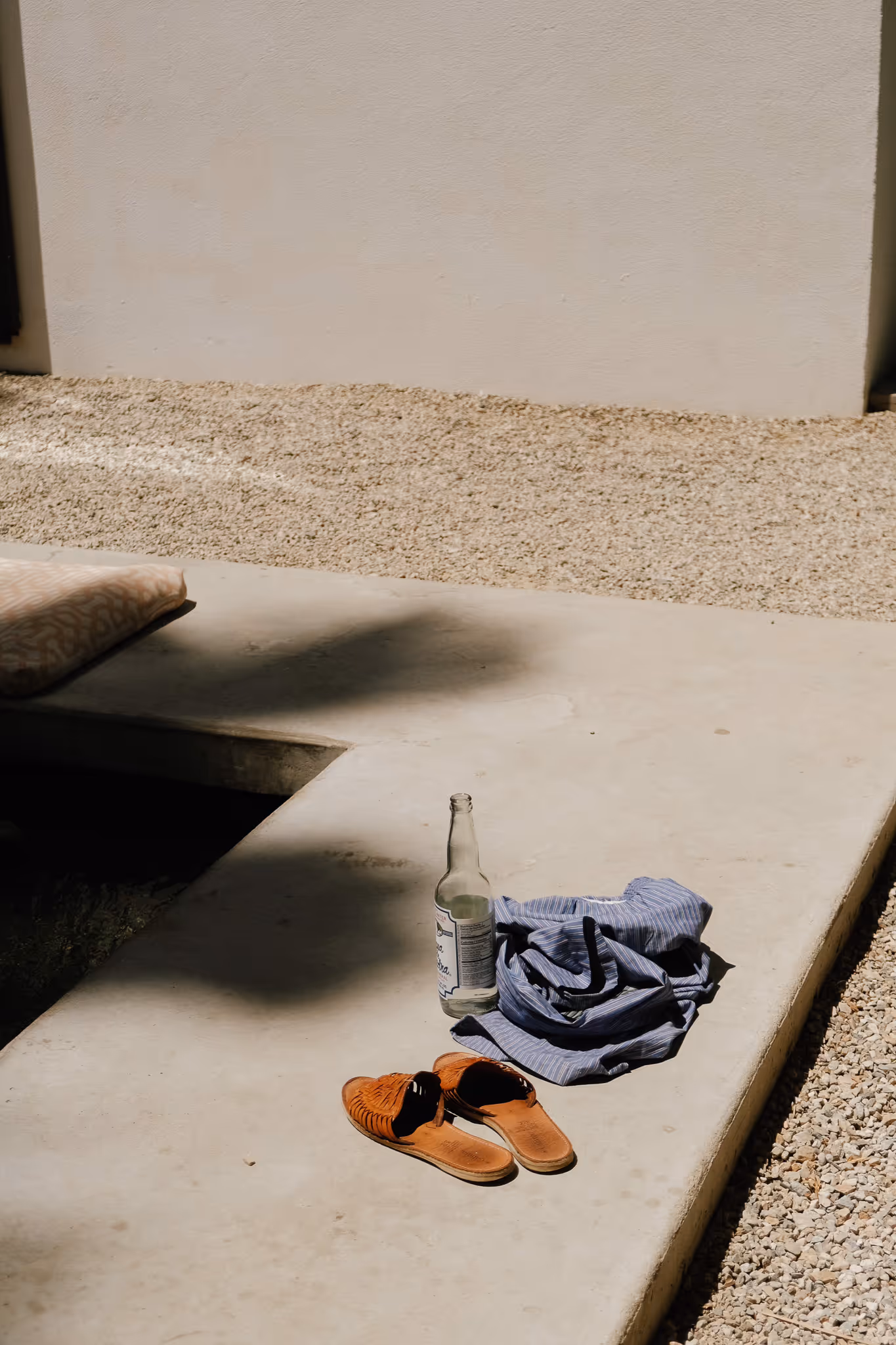 Brown leather sandals, a blue striped shirt, and a clear glass bottle placed on a concrete surface outdoors with gravel in the background.