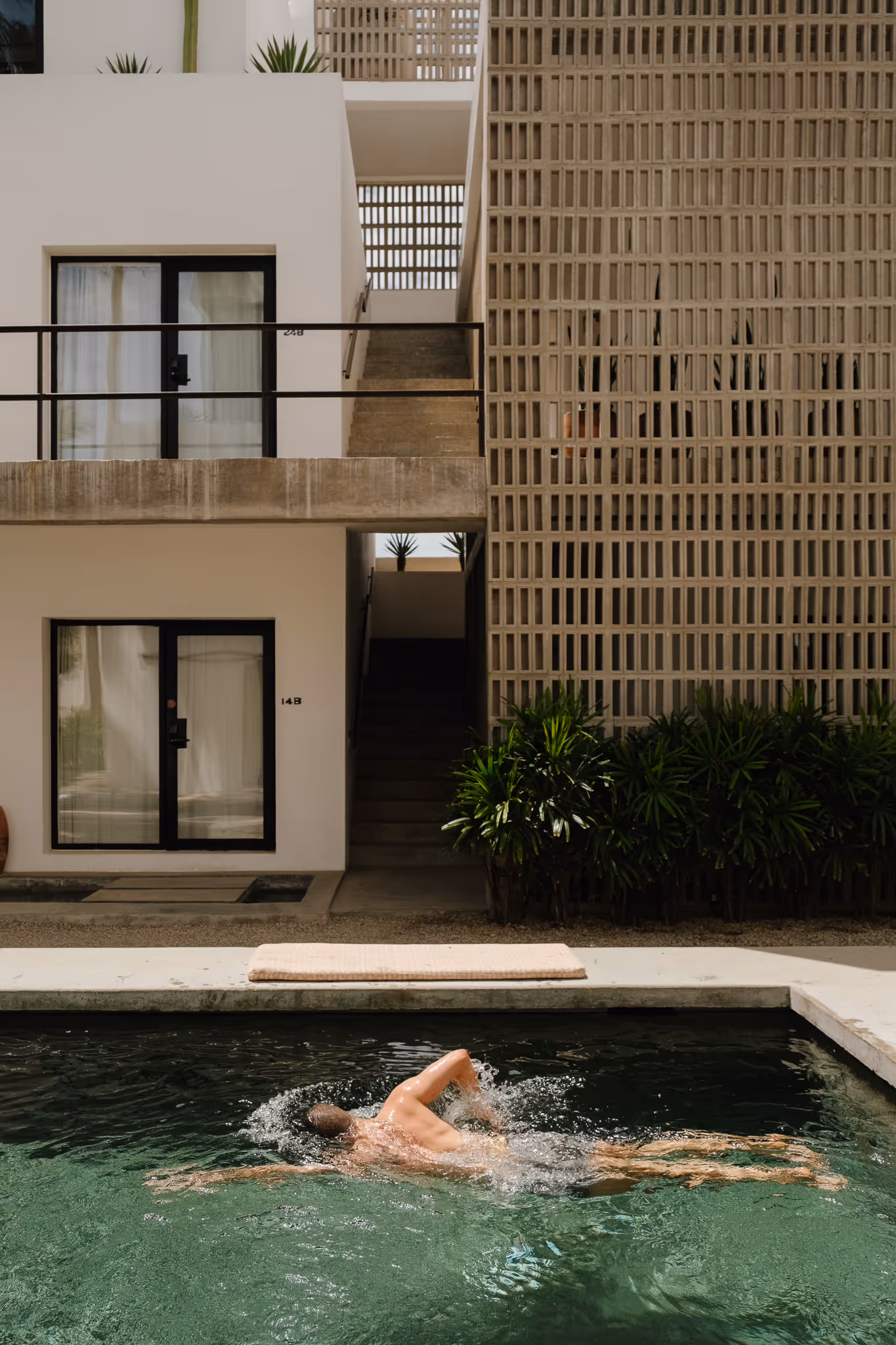 Man swimming freestyle in a pool in front of a modern apartment building with plants and staircases.