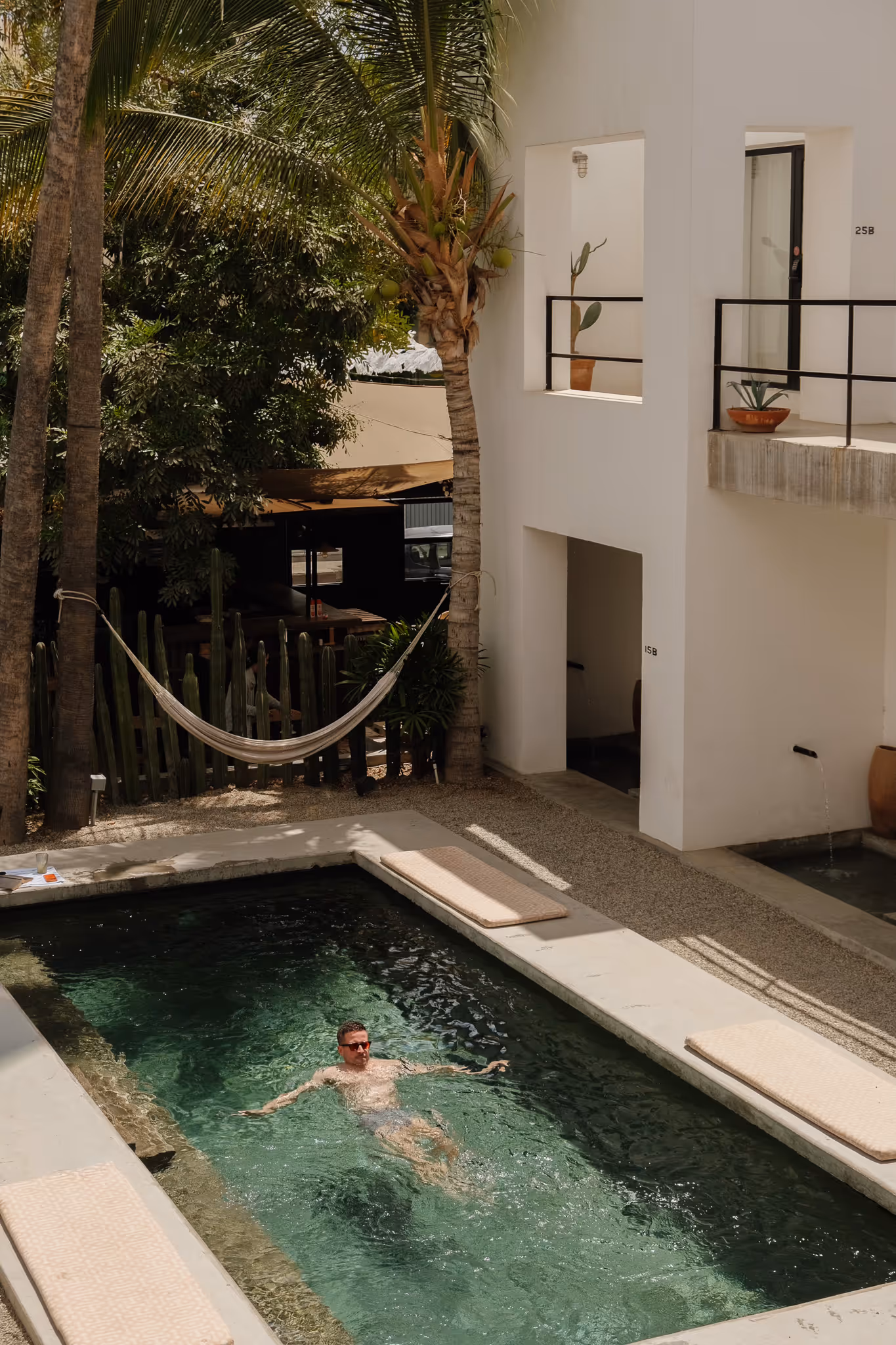 Man wearing sunglasses relaxing and floating in a rectangular outdoor pool surrounded by palm trees and white modern building walls.