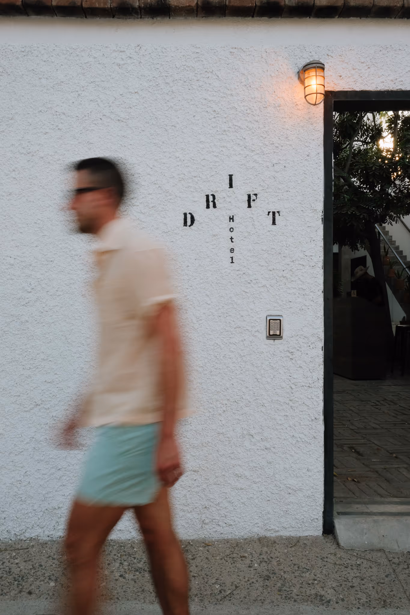 Blurred man walking past a white textured wall with 'DRIFT Hotel' text next to an open door with a glowing outdoor light above it.