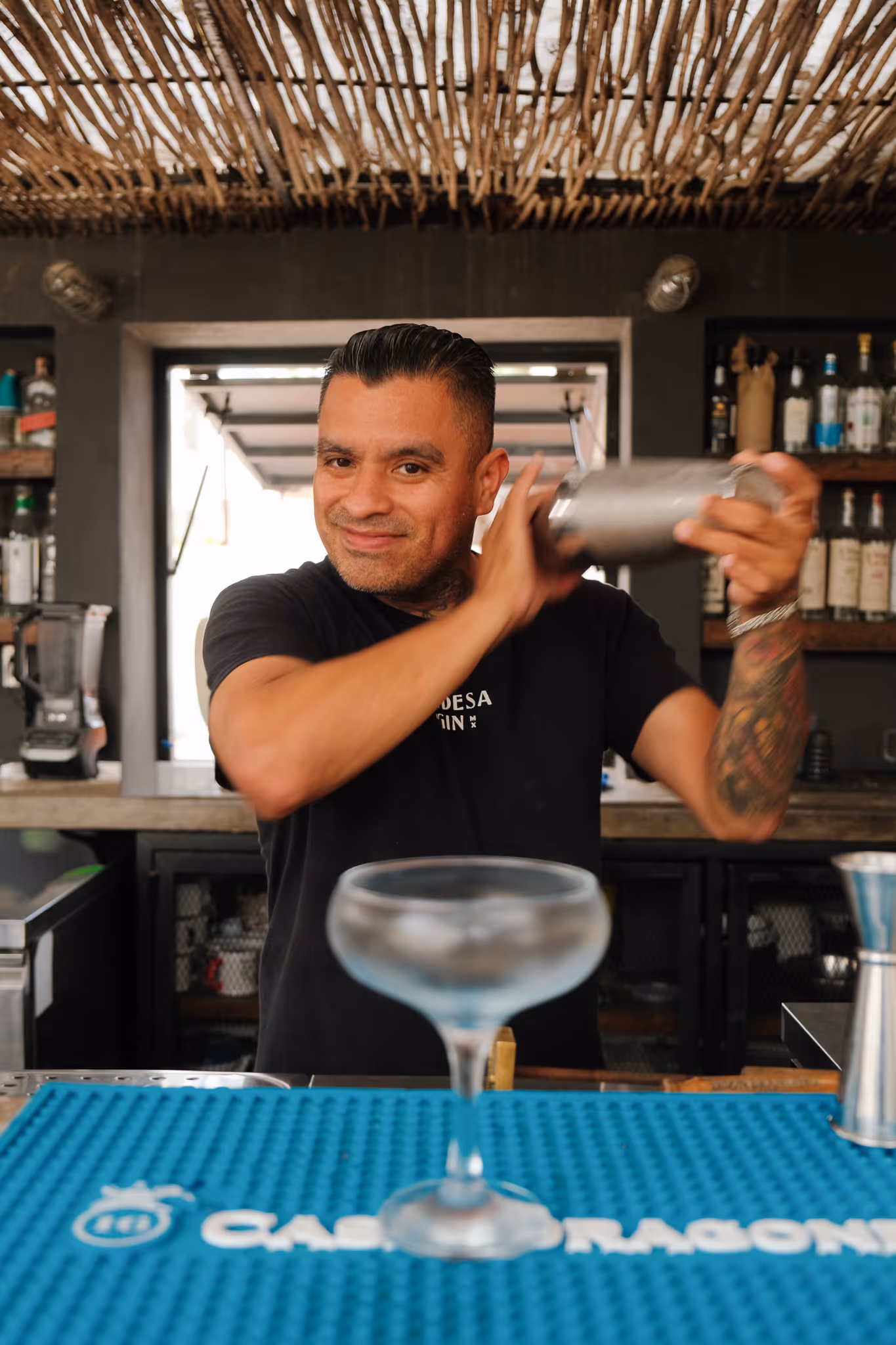 Smiling bartender shaking a cocktail shaker behind a bar with a coupe glass in the foreground.