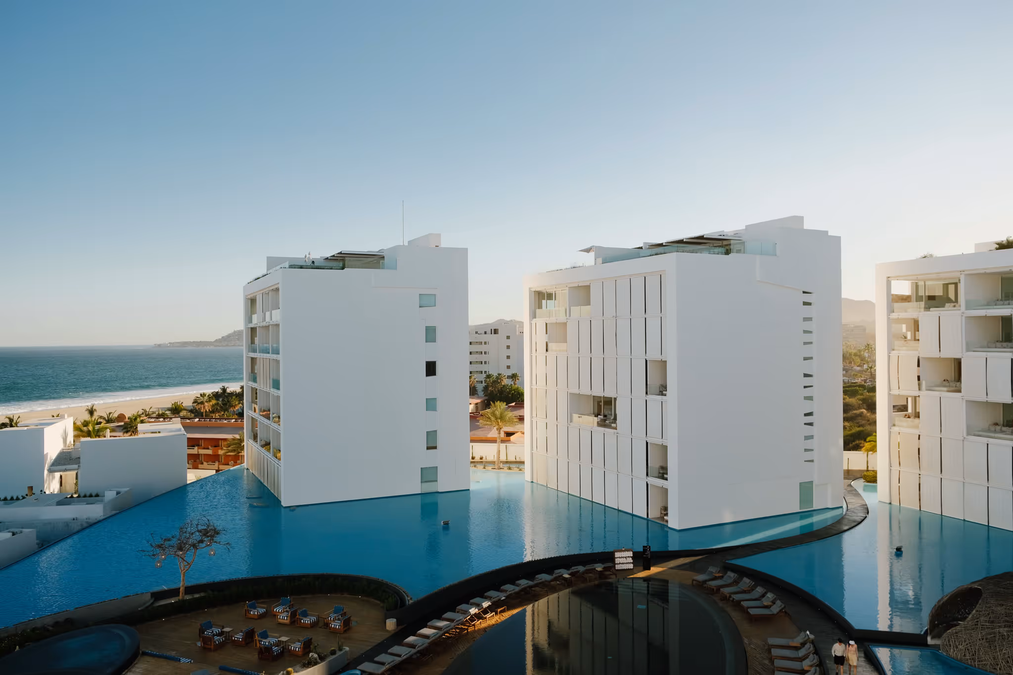 Modern hotel room with large windows overlooking a city skyline, featuring a wooden table, cushioned bench, and a mounted flat-screen TV.
