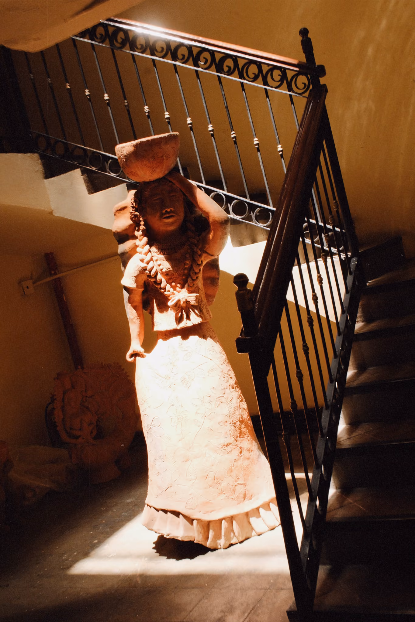 Terracotta statue of a woman carrying a bowl on her head near a staircase with metal railing.