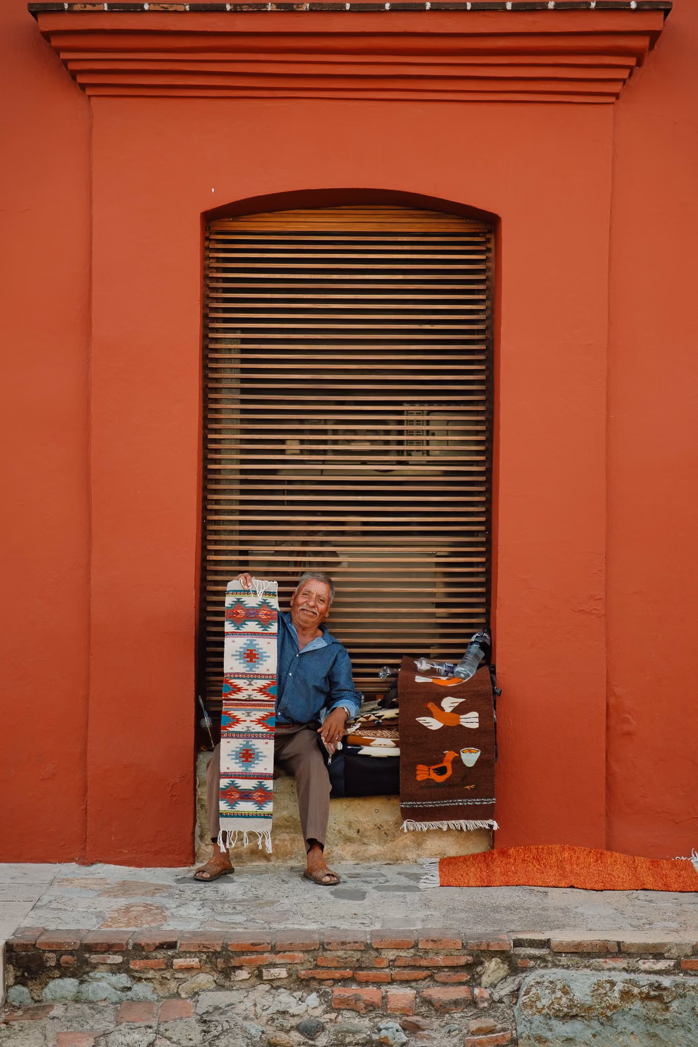 Smiling man sitting in a red wall alcove holding a colorful woven rug and displaying another beside him.