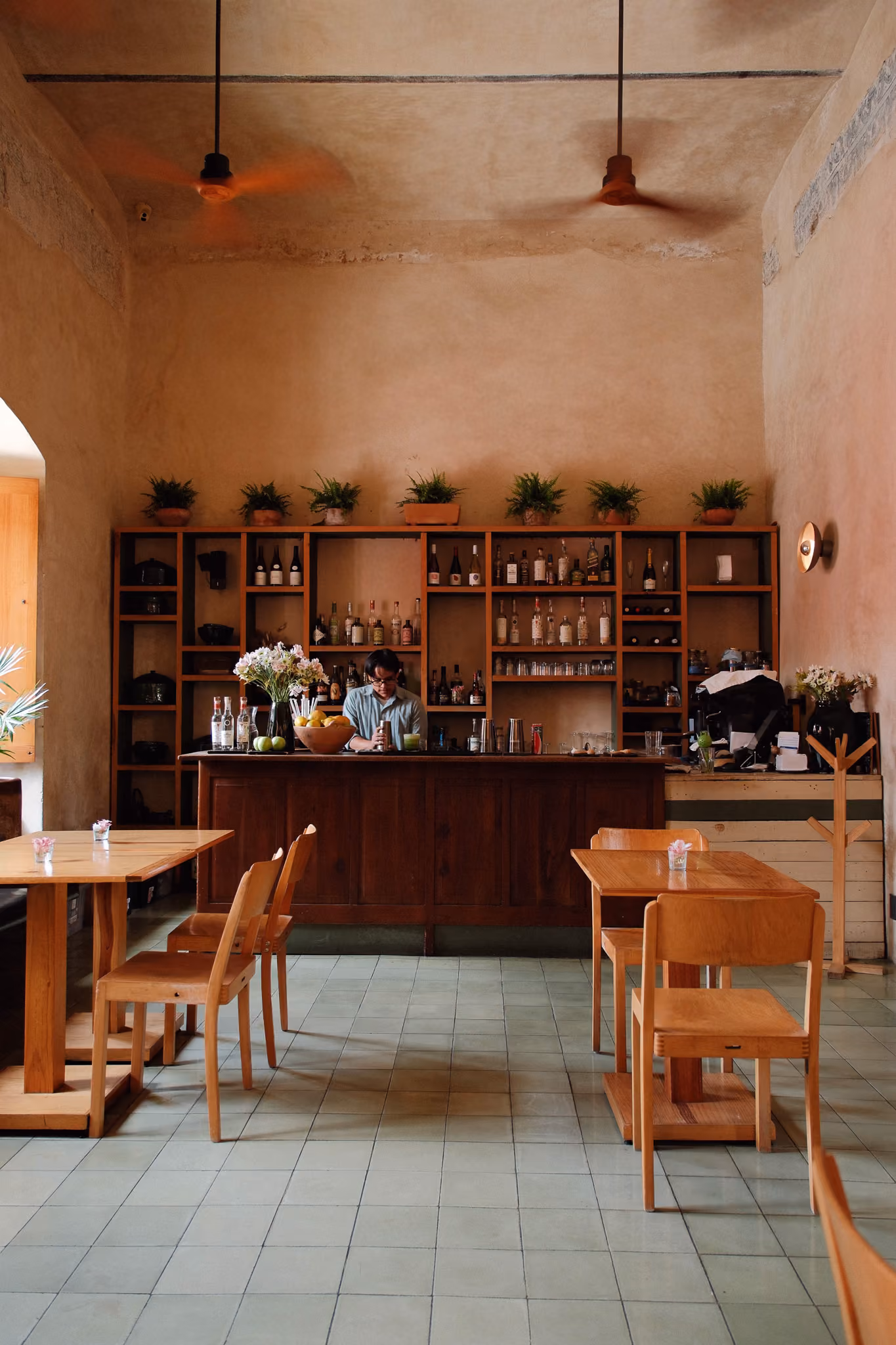 Interior of a cozy bar with wooden tables, chairs, shelves with bottles, and a bartender behind the counter.
