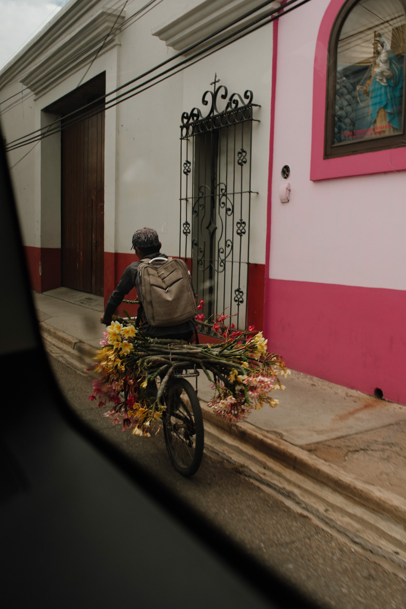 Older man wearing a mask riding a bicycle on a city sidewalk with buildings and vehicles in the background.