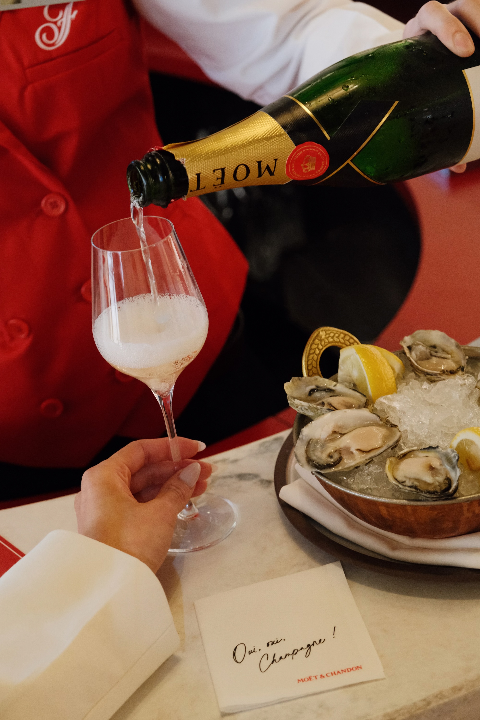 A person pours Moët champagne into a glass held by another over a table with oysters on ice.