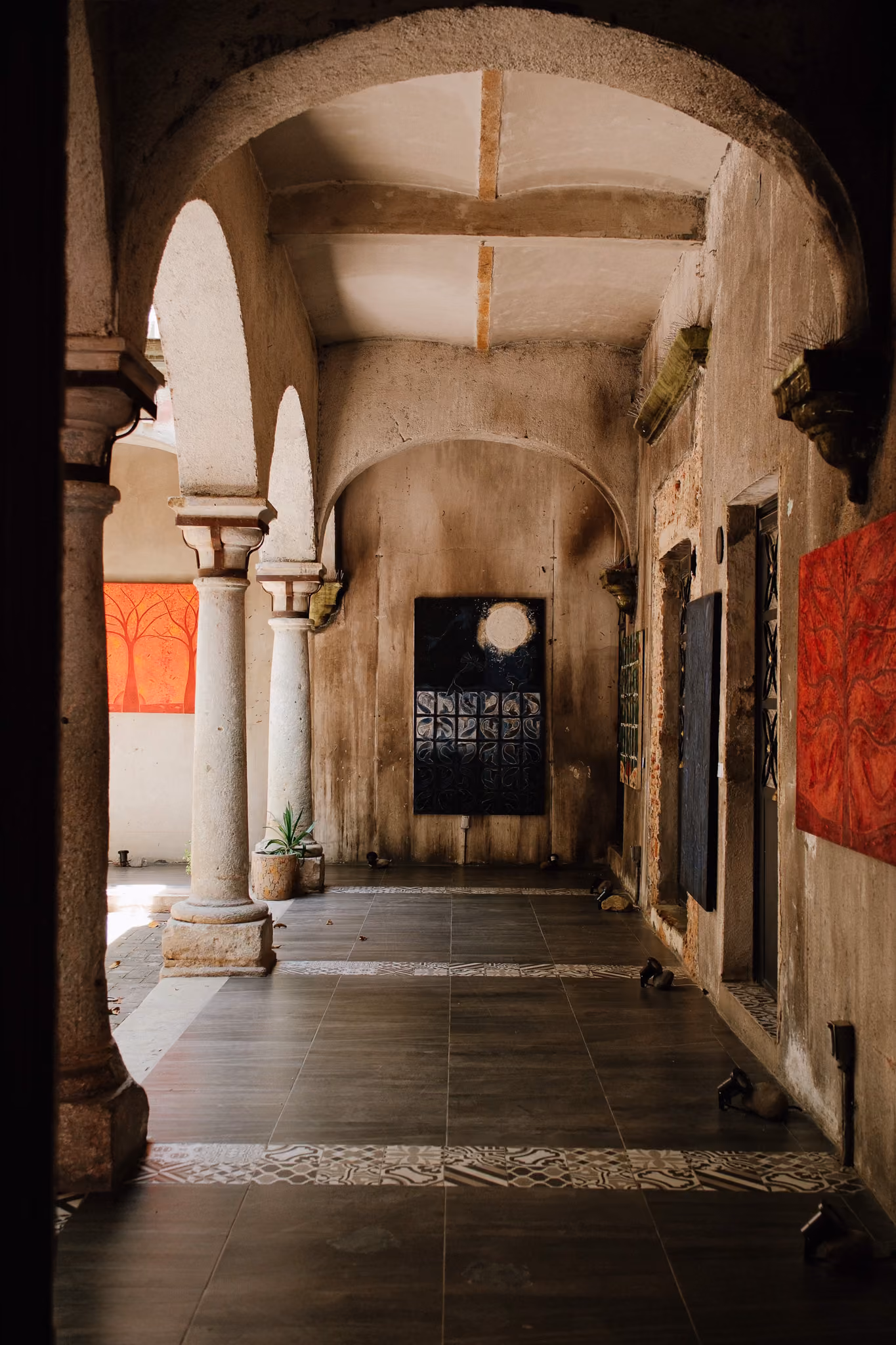 Arched stone corridor with tiled floor, potted plants, and abstract paintings on textured walls.
