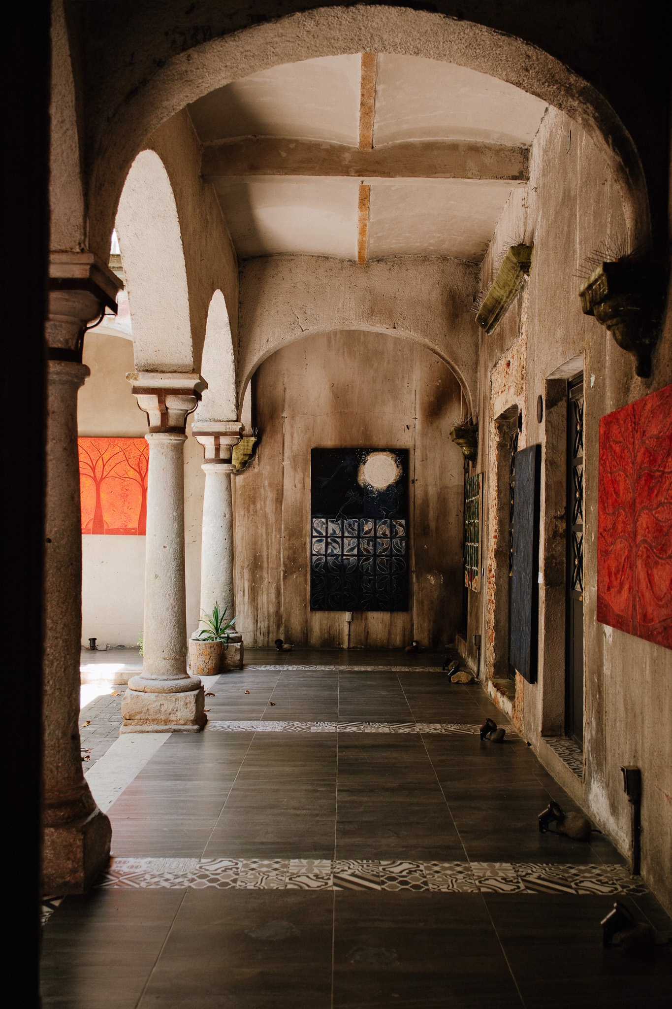 Arched stone corridor with tiled floor, potted plants, and abstract paintings on textured walls.