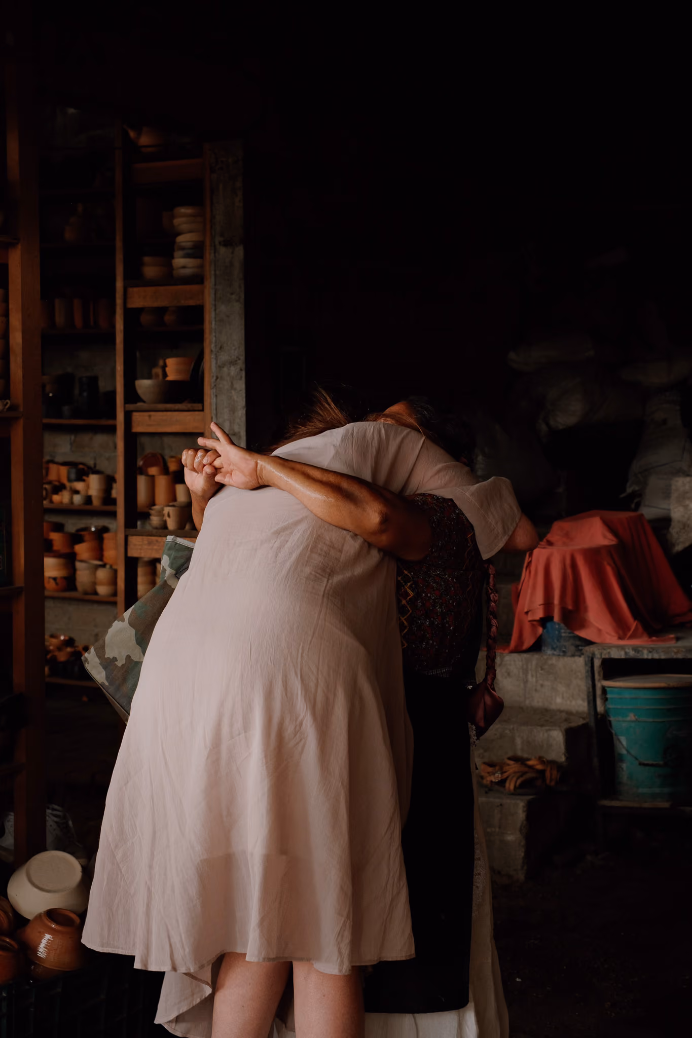 Two women embrace warmly in a pottery workshop filled with clay pots and shelves.