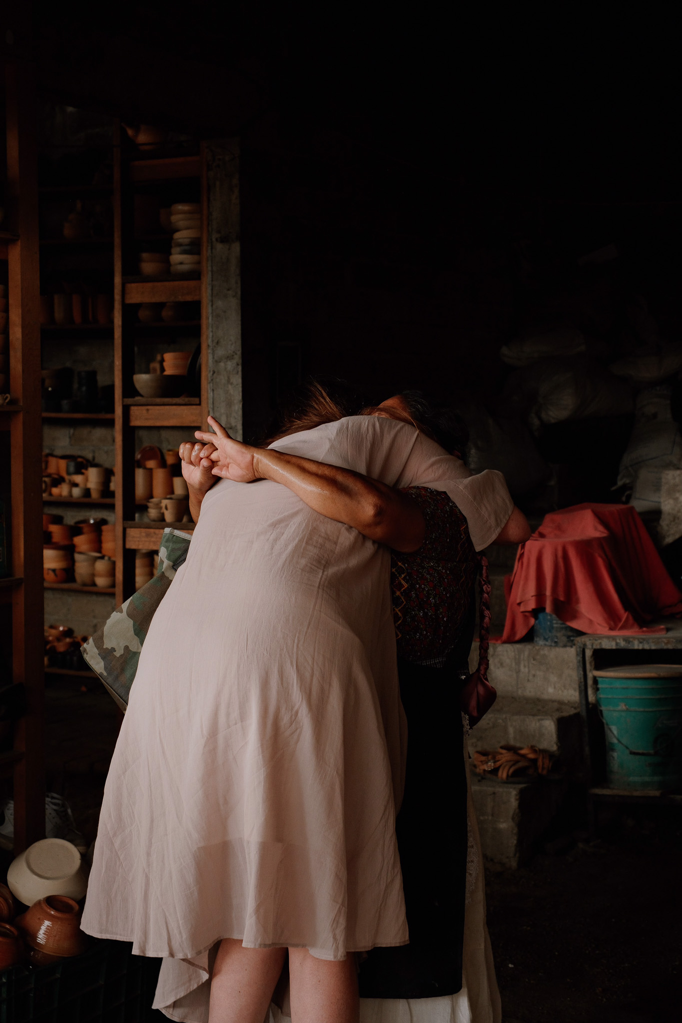 Two women embrace warmly in a pottery workshop filled with clay pots and shelves.
