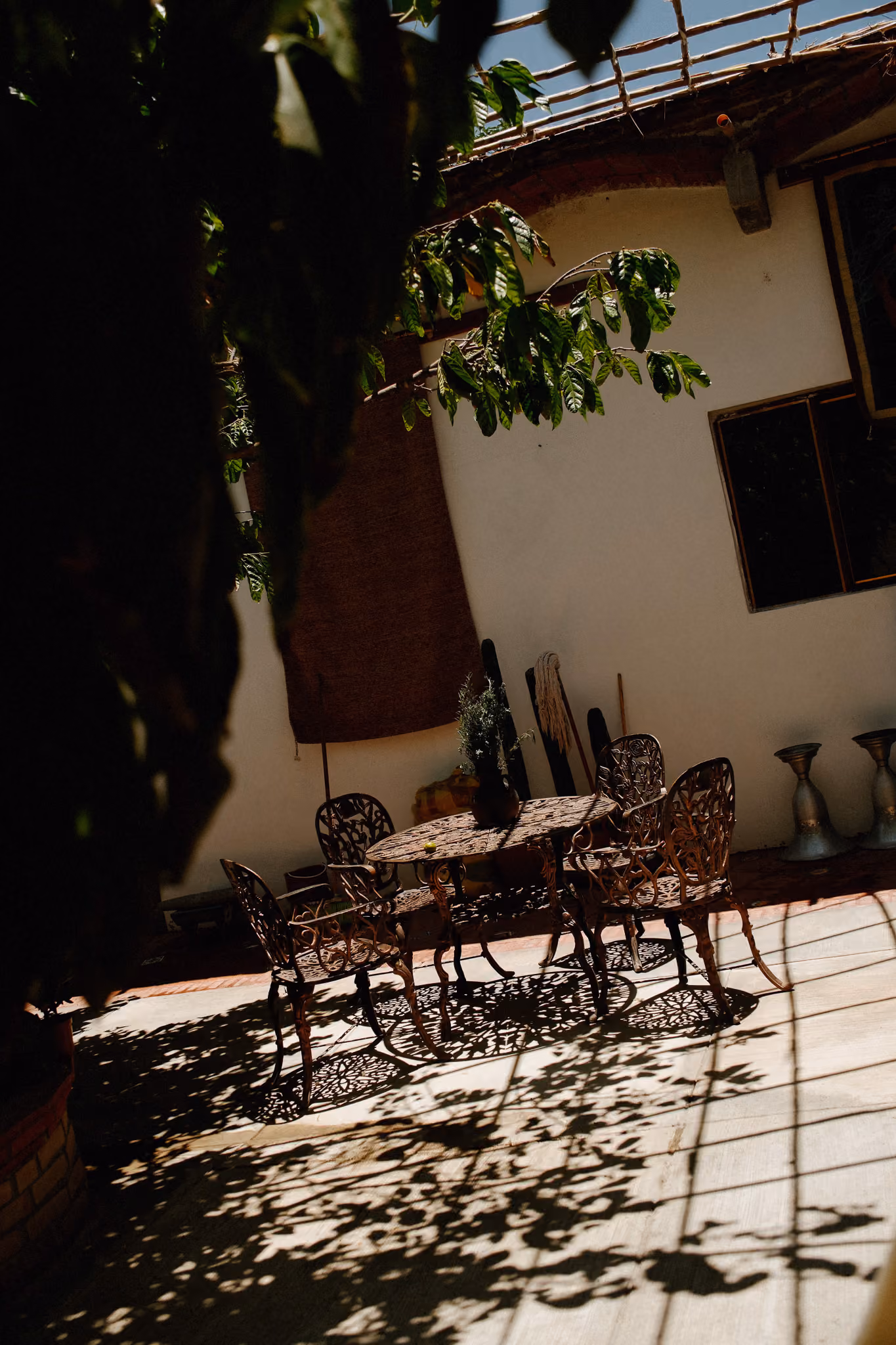 Iron outdoor table with chairs casting leaf-patterned shadows on a sunlit patio under greenery.