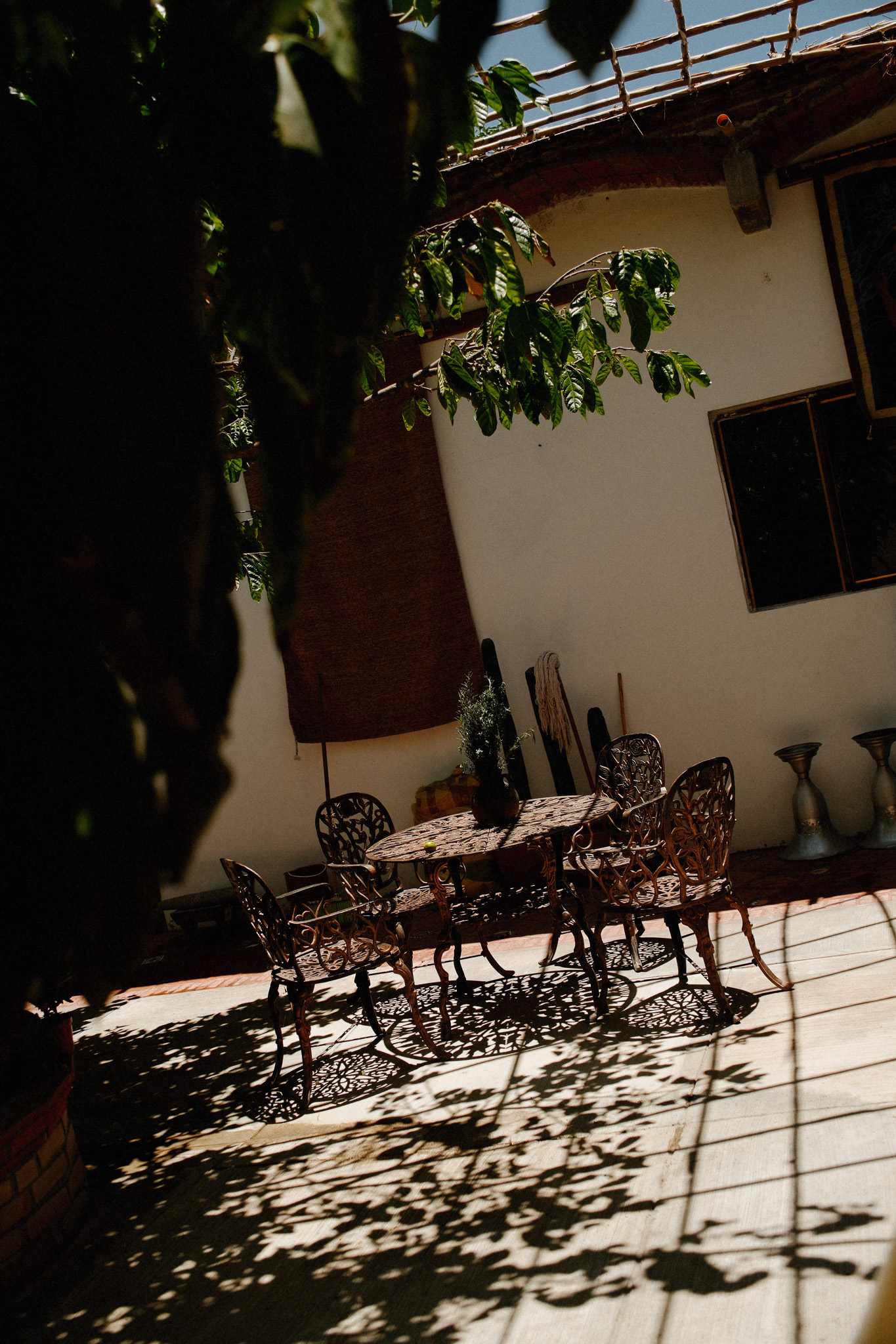 Iron outdoor table with chairs casting leaf-patterned shadows on a sunlit patio under greenery.