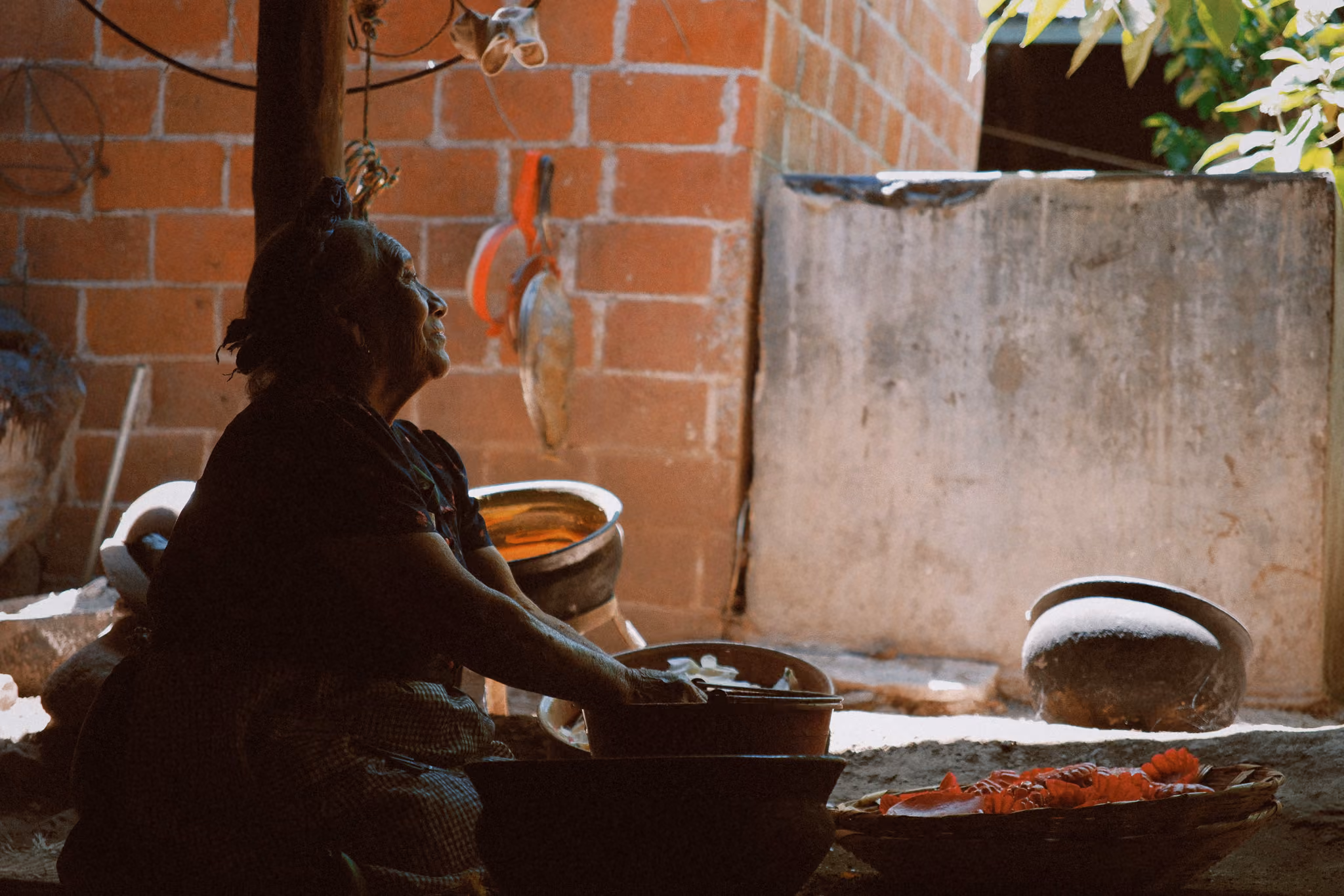 Elderly woman sitting indoors washing or preparing food with clay pots and a basket nearby.