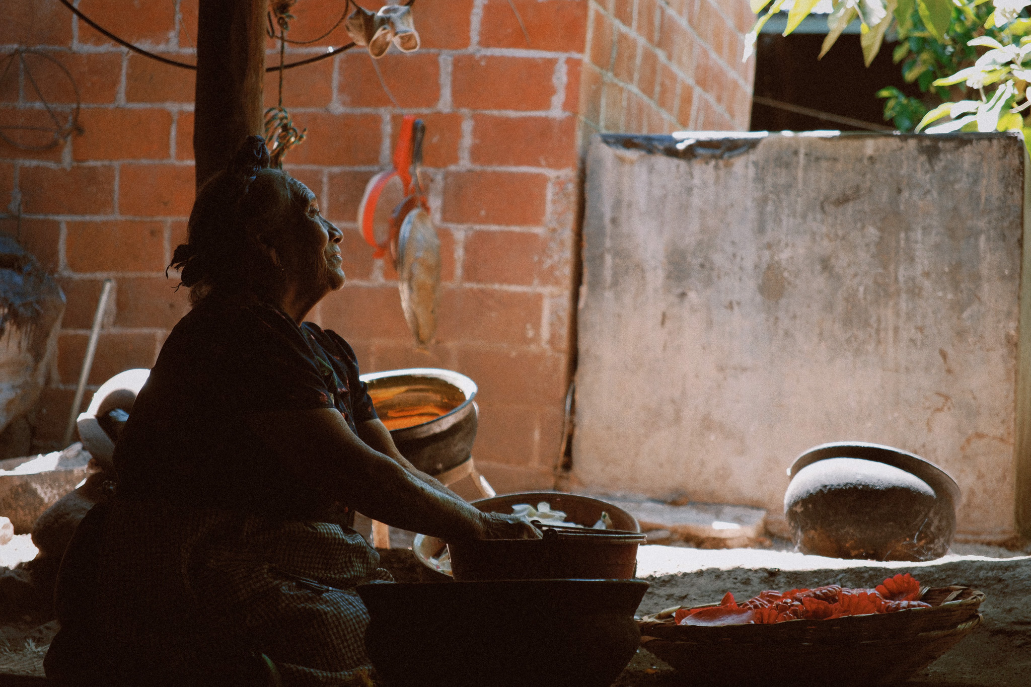 Elderly woman sitting indoors washing or preparing food with clay pots and a basket nearby.