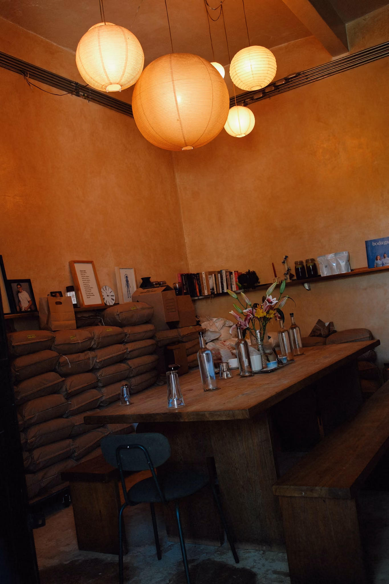Rustic room with a wooden table, glass bottles, flowers, stacked sacks, and hanging paper lanterns.