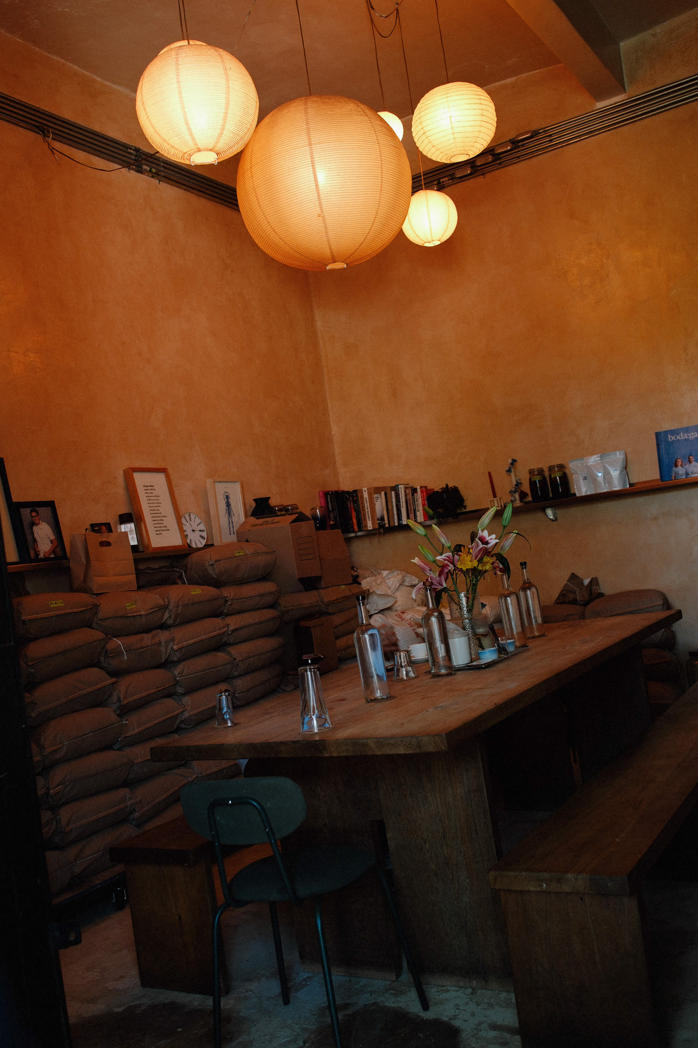 Rustic room with a wooden table, glass bottles, flowers, stacked sacks, and hanging paper lanterns.