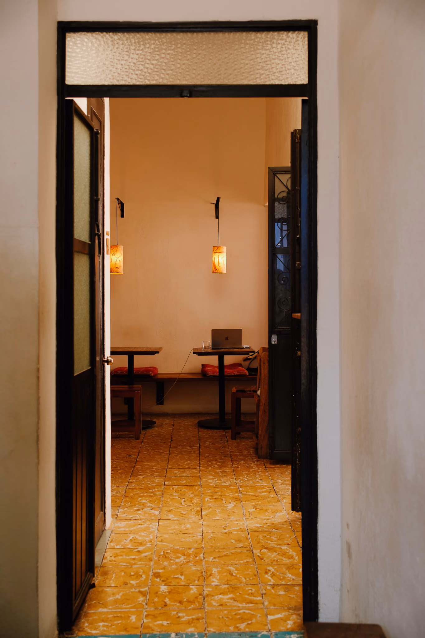 View through doorway into a cozy room with warm lighting, tables, chairs, and a laptop on one table.