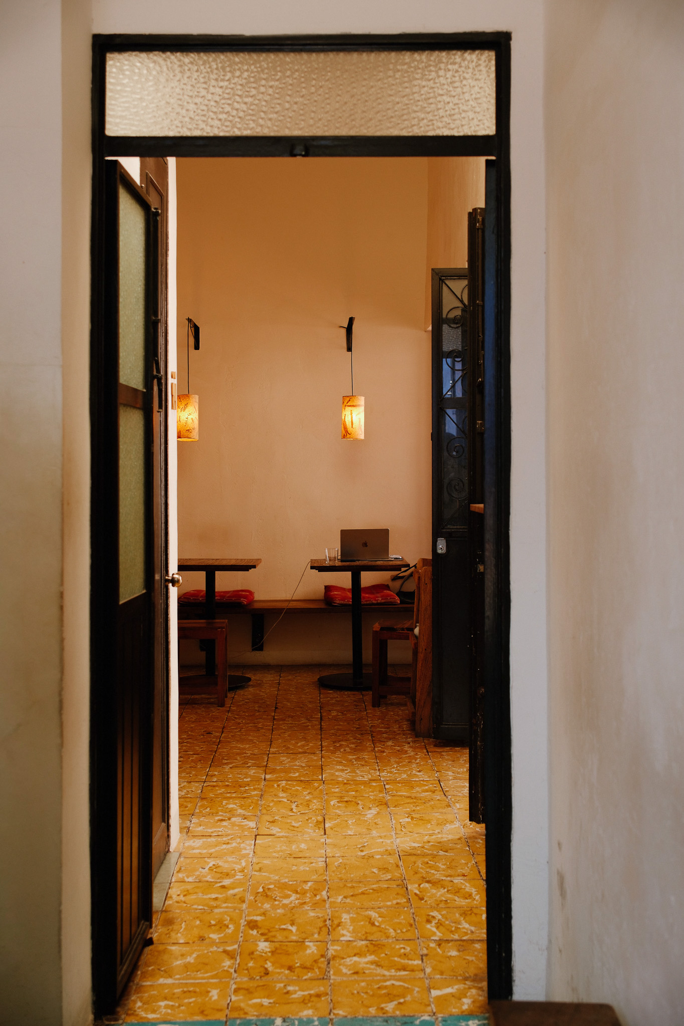 View through doorway into a cozy room with warm lighting, tables, chairs, and a laptop on one table.