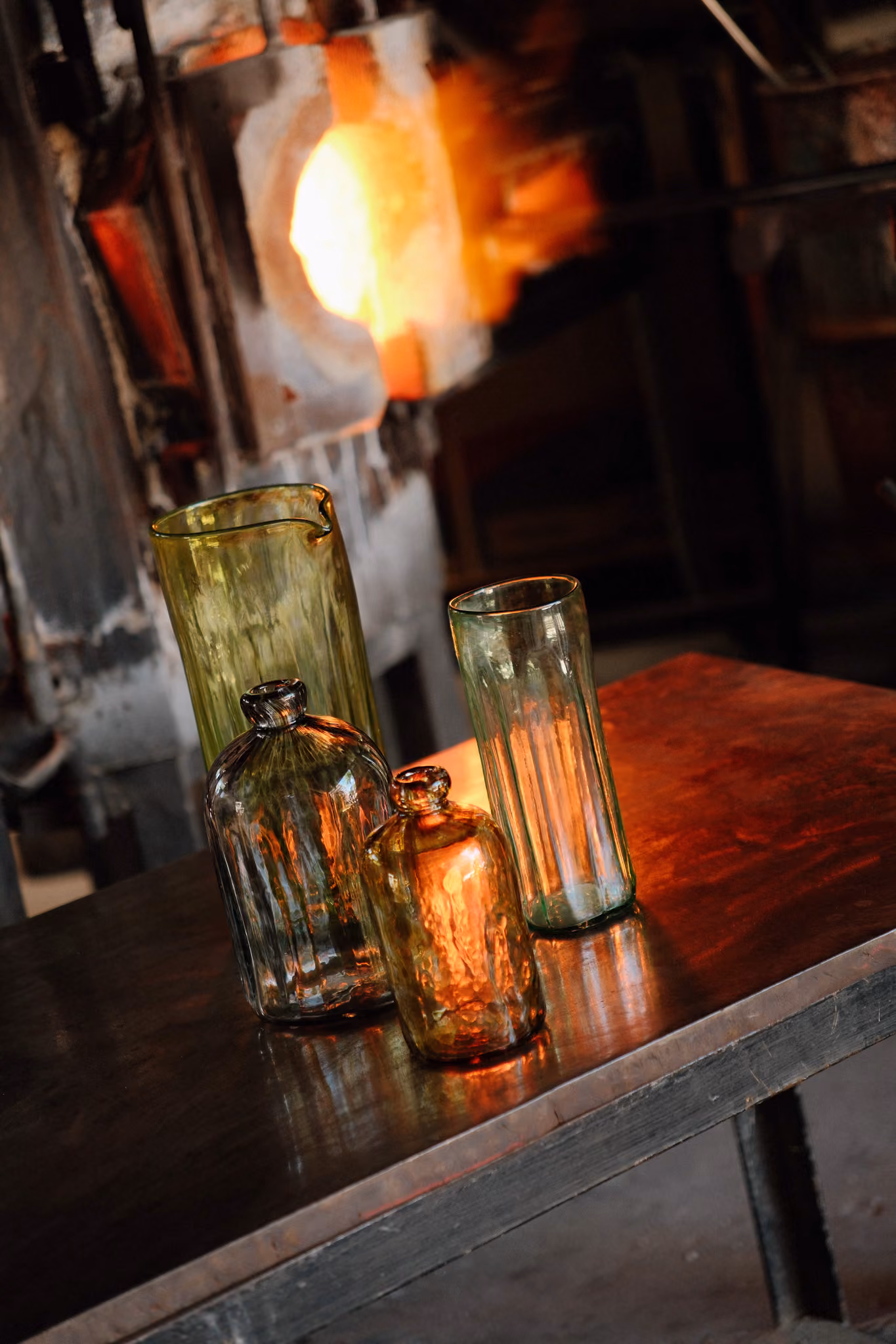 Four handcrafted glass bottles and pitchers on a table near a glowing glass furnace.