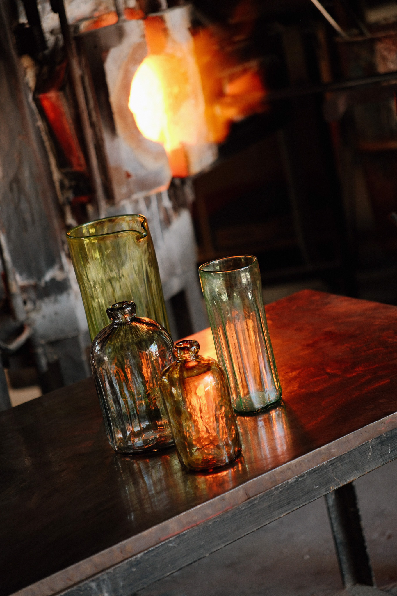 Four handcrafted glass bottles and pitchers on a table near a glowing glass furnace.