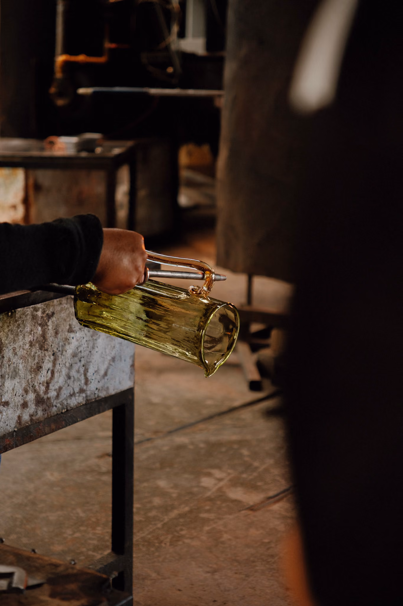 Hand holding a tool shaping a yellow glass pitcher in a glassblowing workshop.