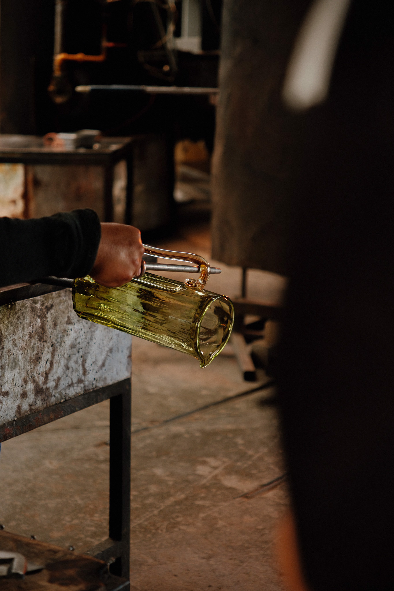 Hand holding a tool shaping a yellow glass pitcher in a glassblowing workshop.