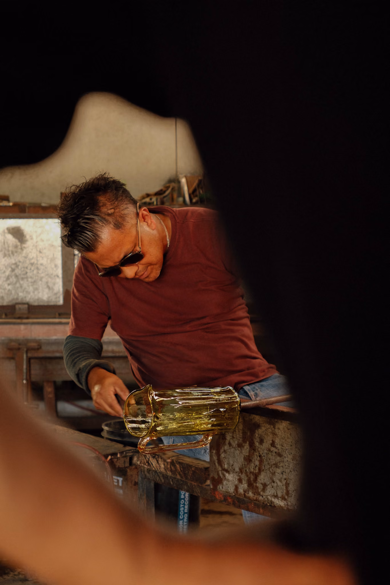 Man wearing sunglasses shaping molten glass on a metal bench in a glassblowing workshop.
