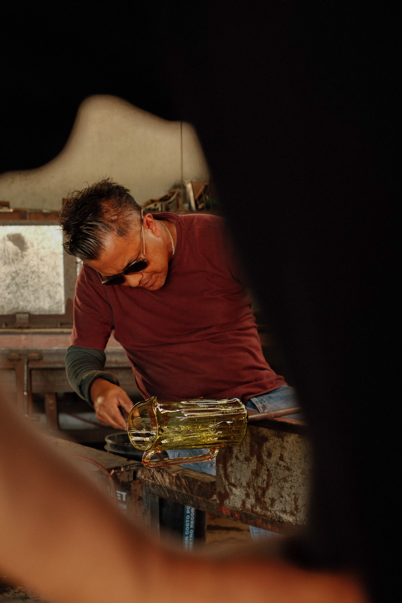 Man wearing sunglasses shaping molten glass on a metal bench in a glassblowing workshop.