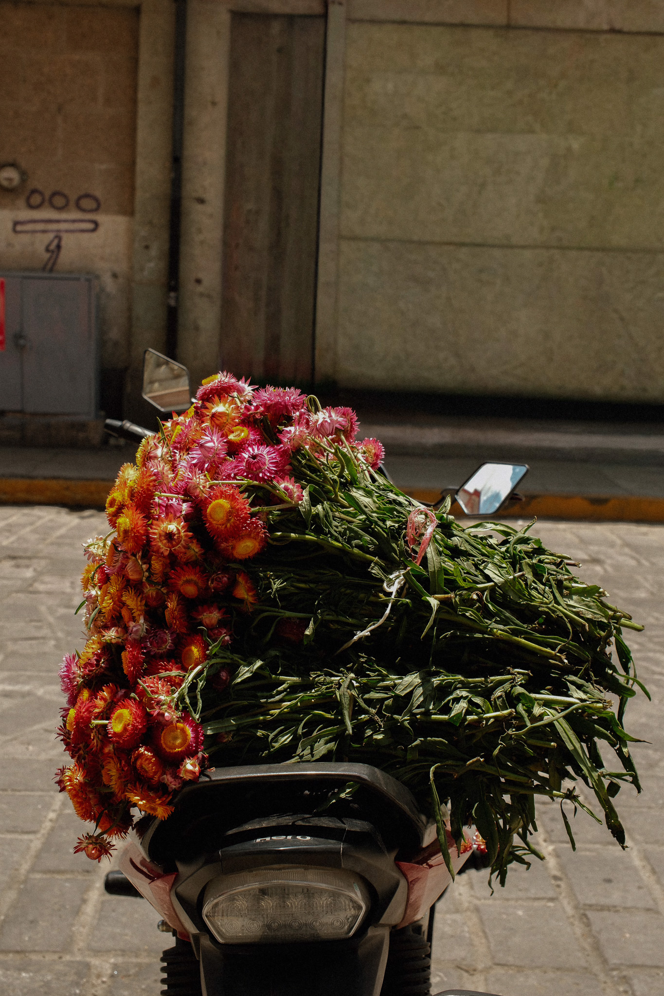 Bundles of bright pink and orange flowers tied and placed on the back of a parked motorcycle.