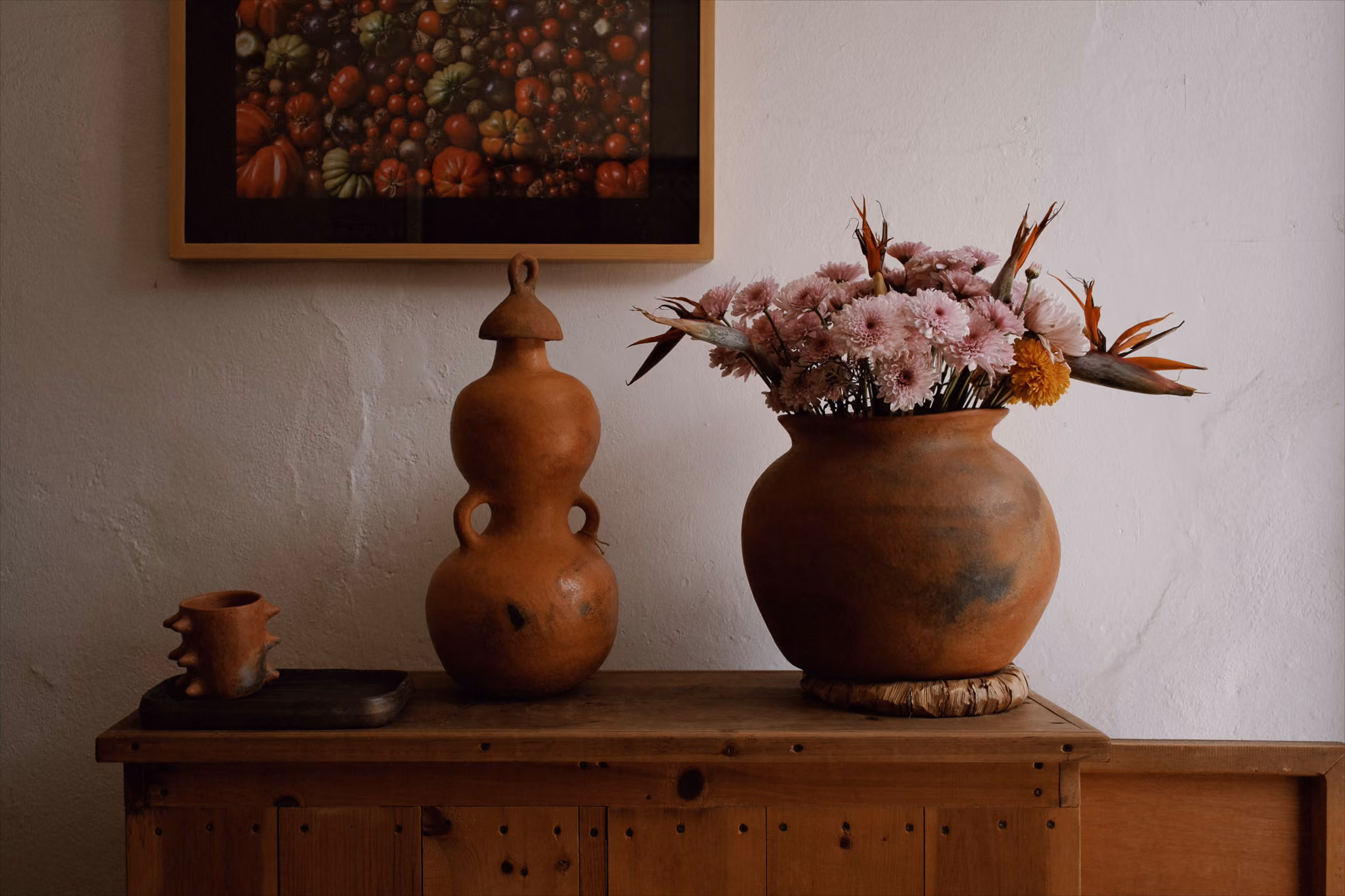 Rustic wooden table with terracotta vase holding pink flowers and decorative pottery on a white wall.