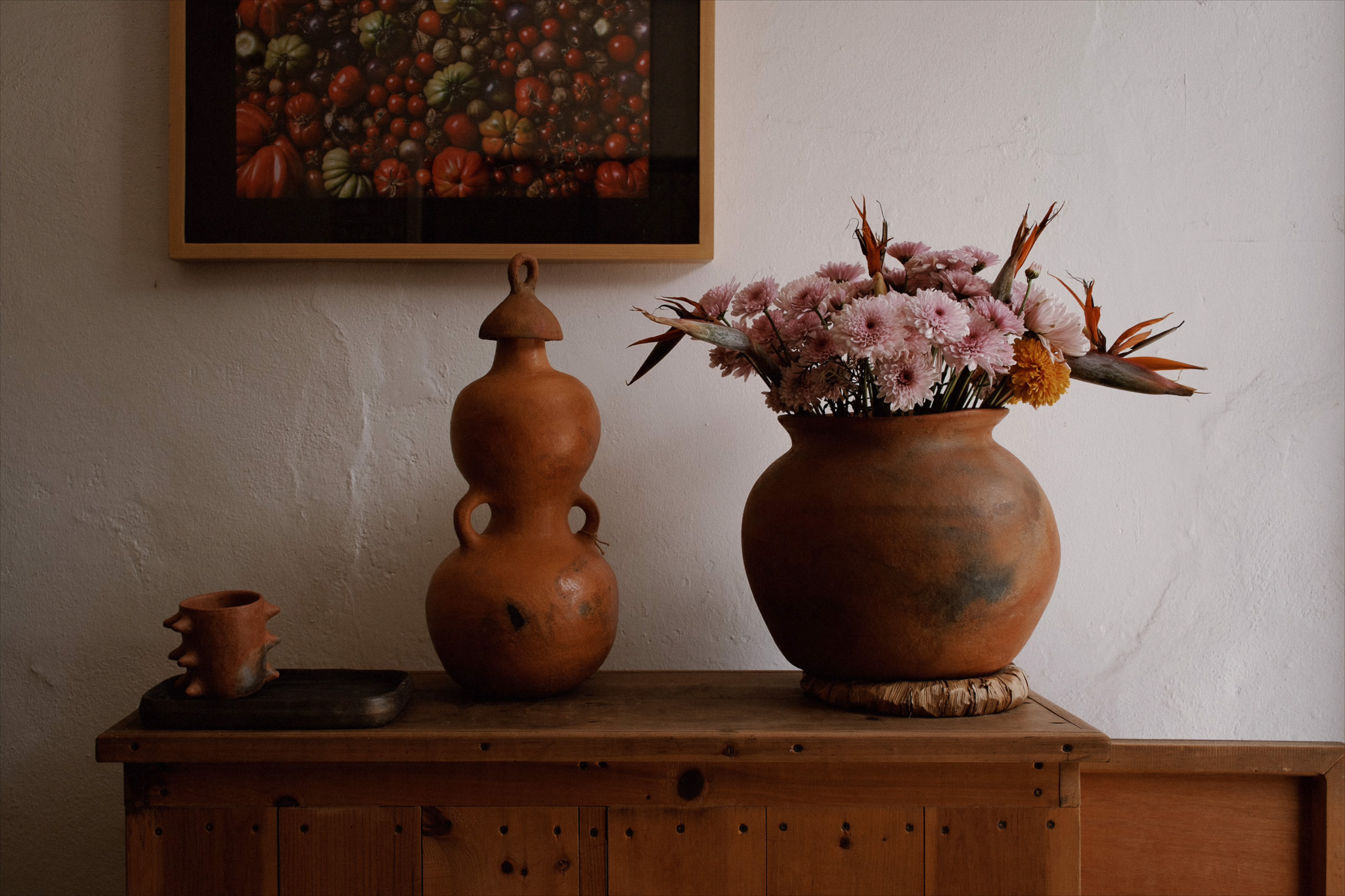 Rustic wooden table with terracotta vase holding pink flowers and decorative pottery on a white wall.