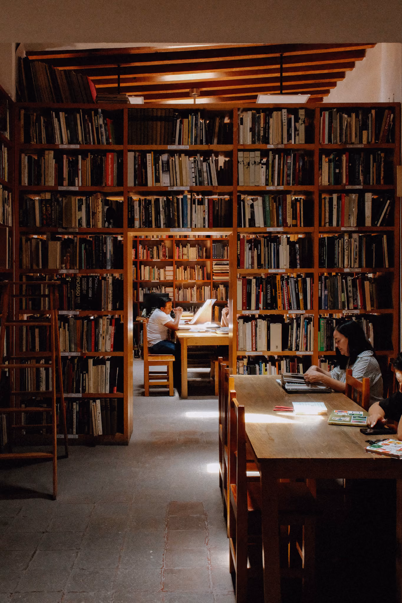 People sitting at wooden tables reading in a warmly lit library with tall bookshelves.