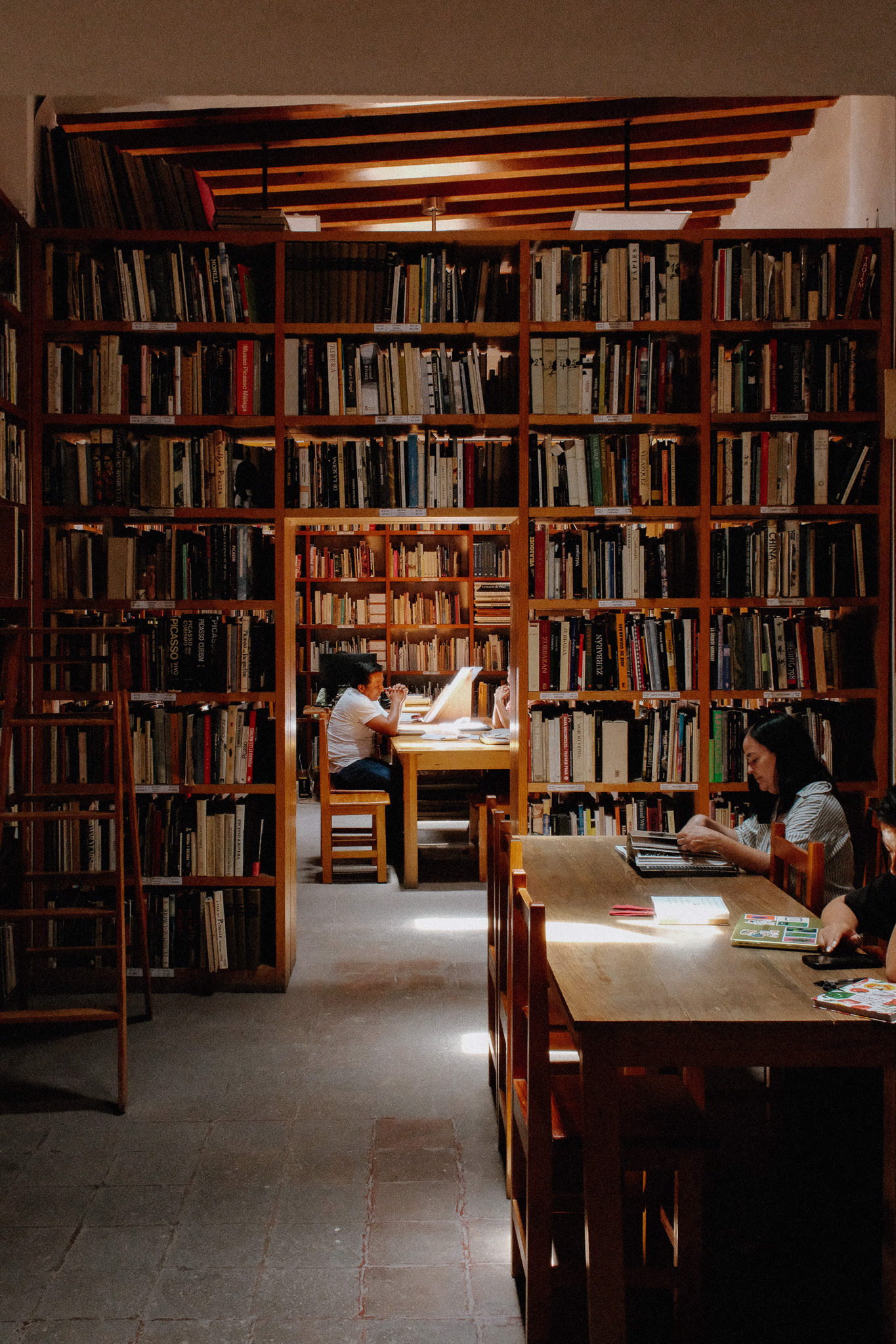 People sitting at wooden tables reading in a warmly lit library with tall bookshelves.