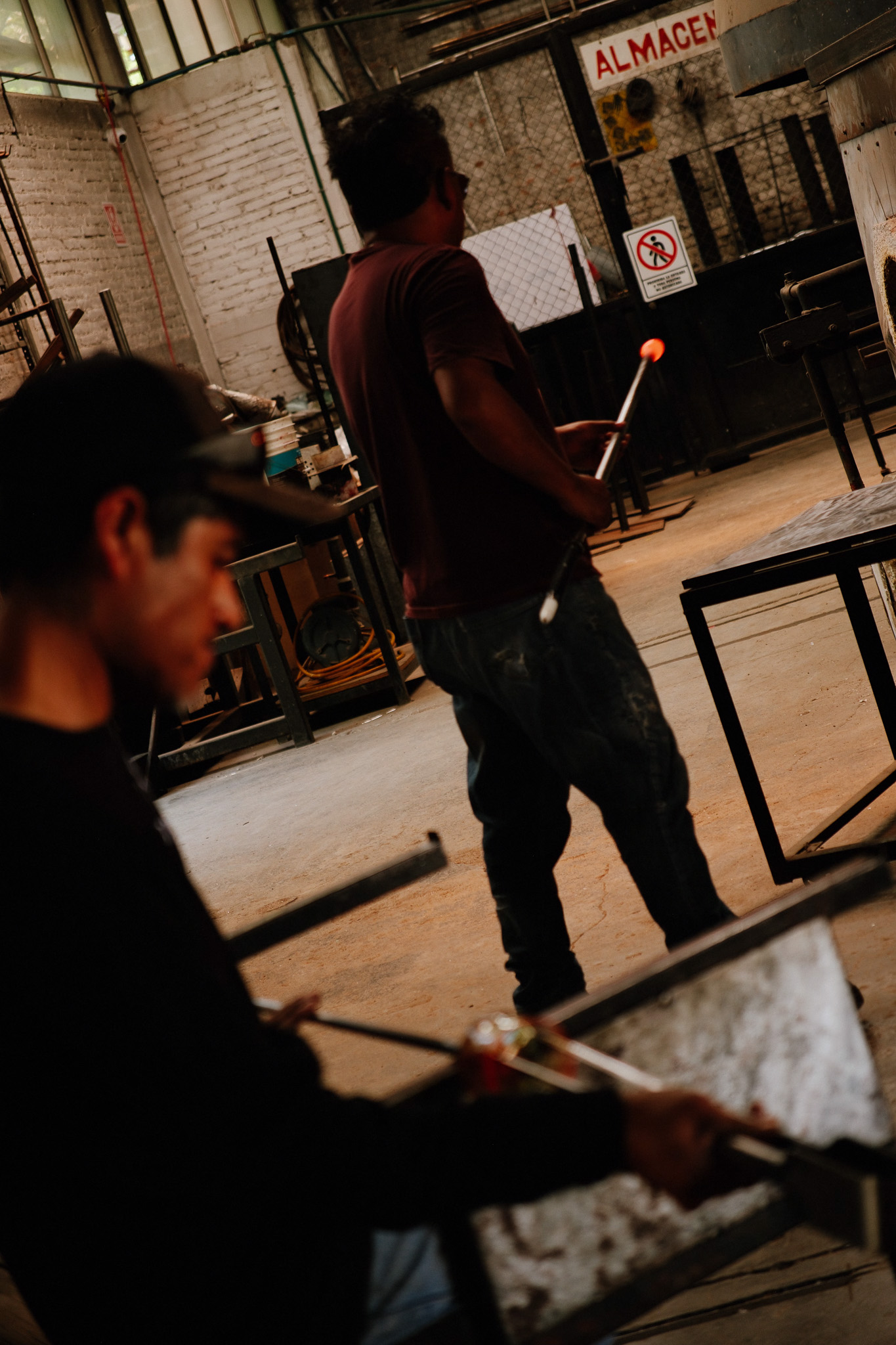 Two men working in a workshop, one holding a glowing glass rod, making glass objects.