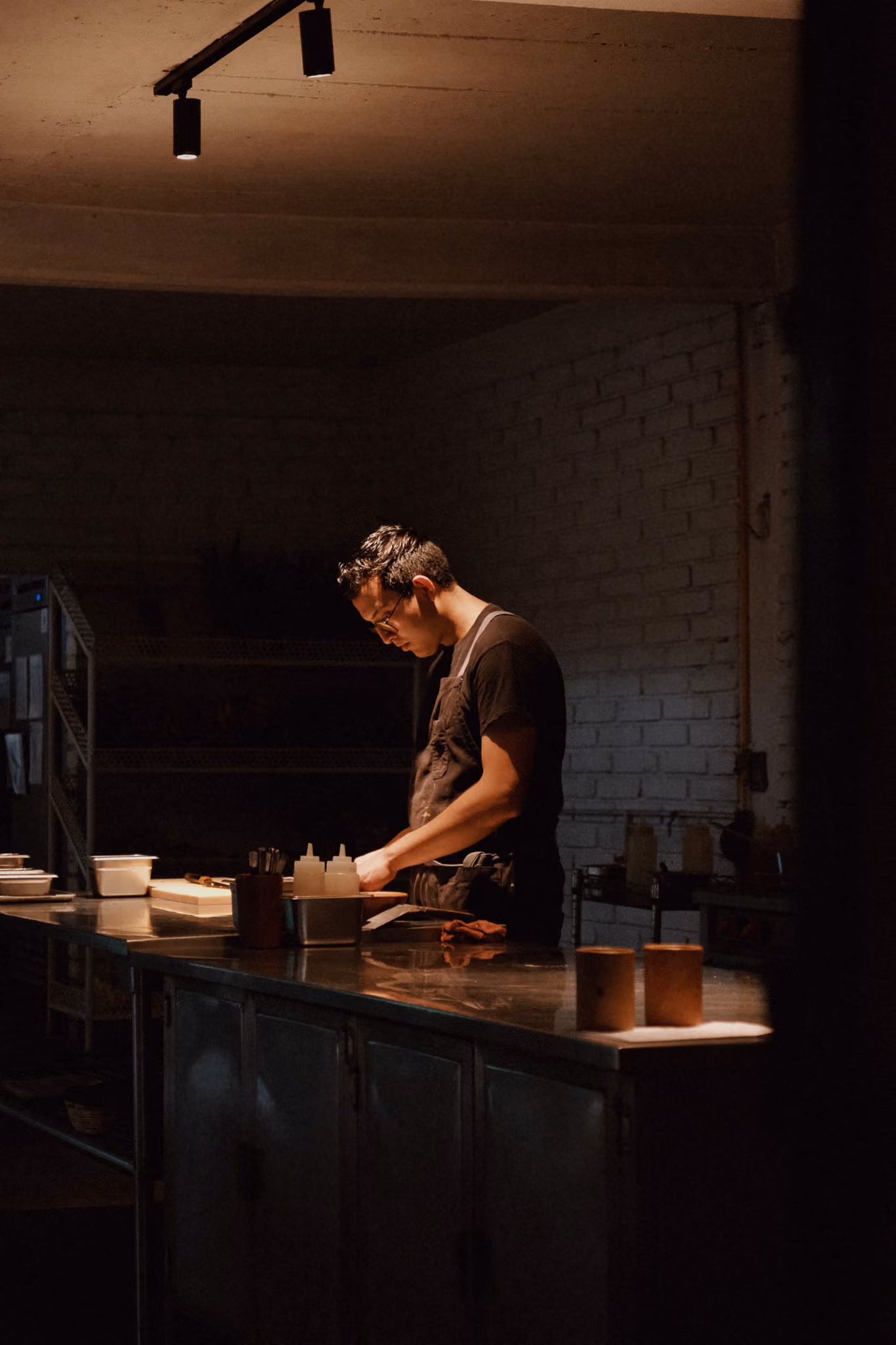 Man in apron working on food preparation at a dimly lit kitchen counter.