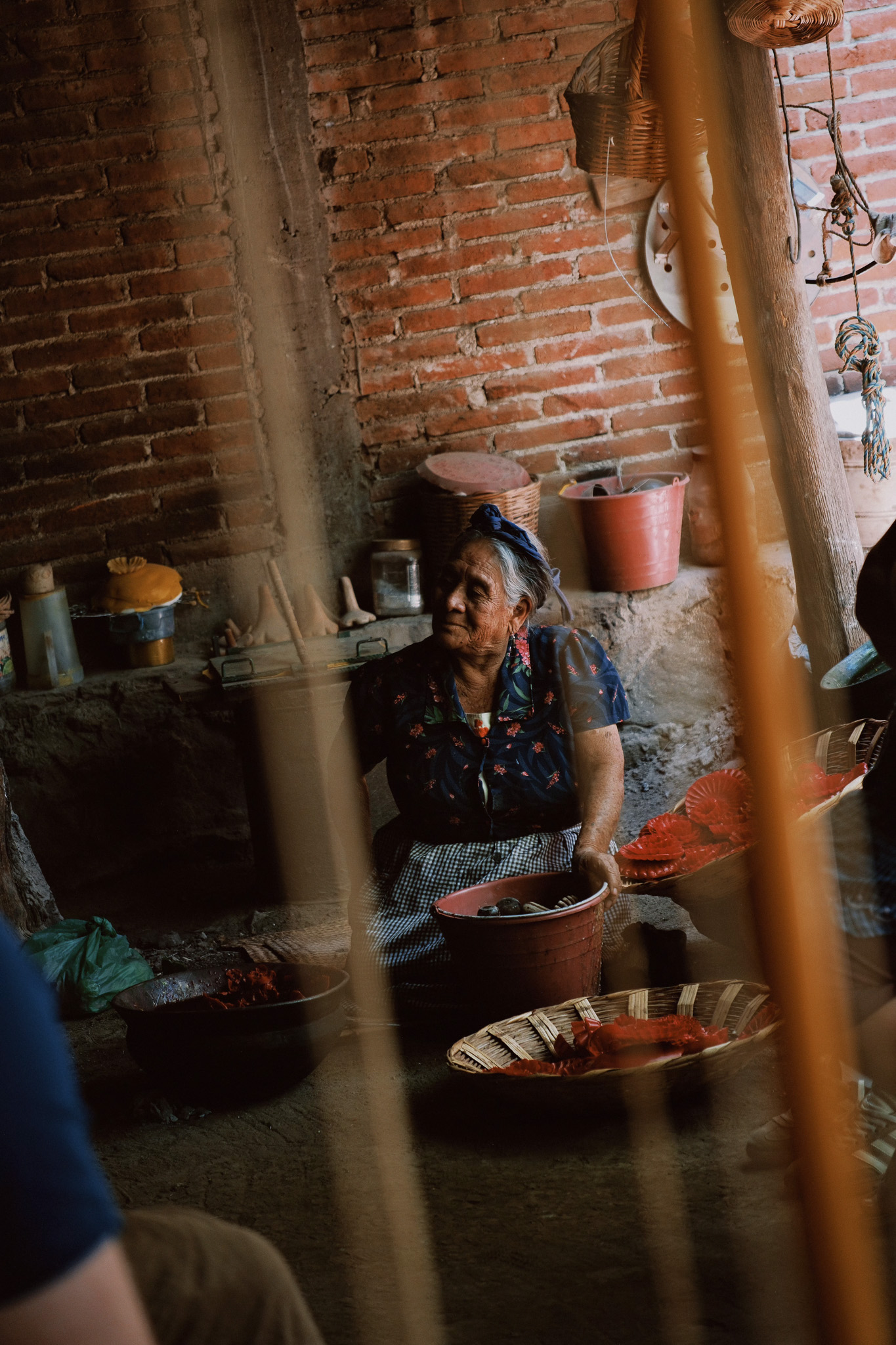 Elderly woman sitting indoors holding a red basket with pots and baskets around her.