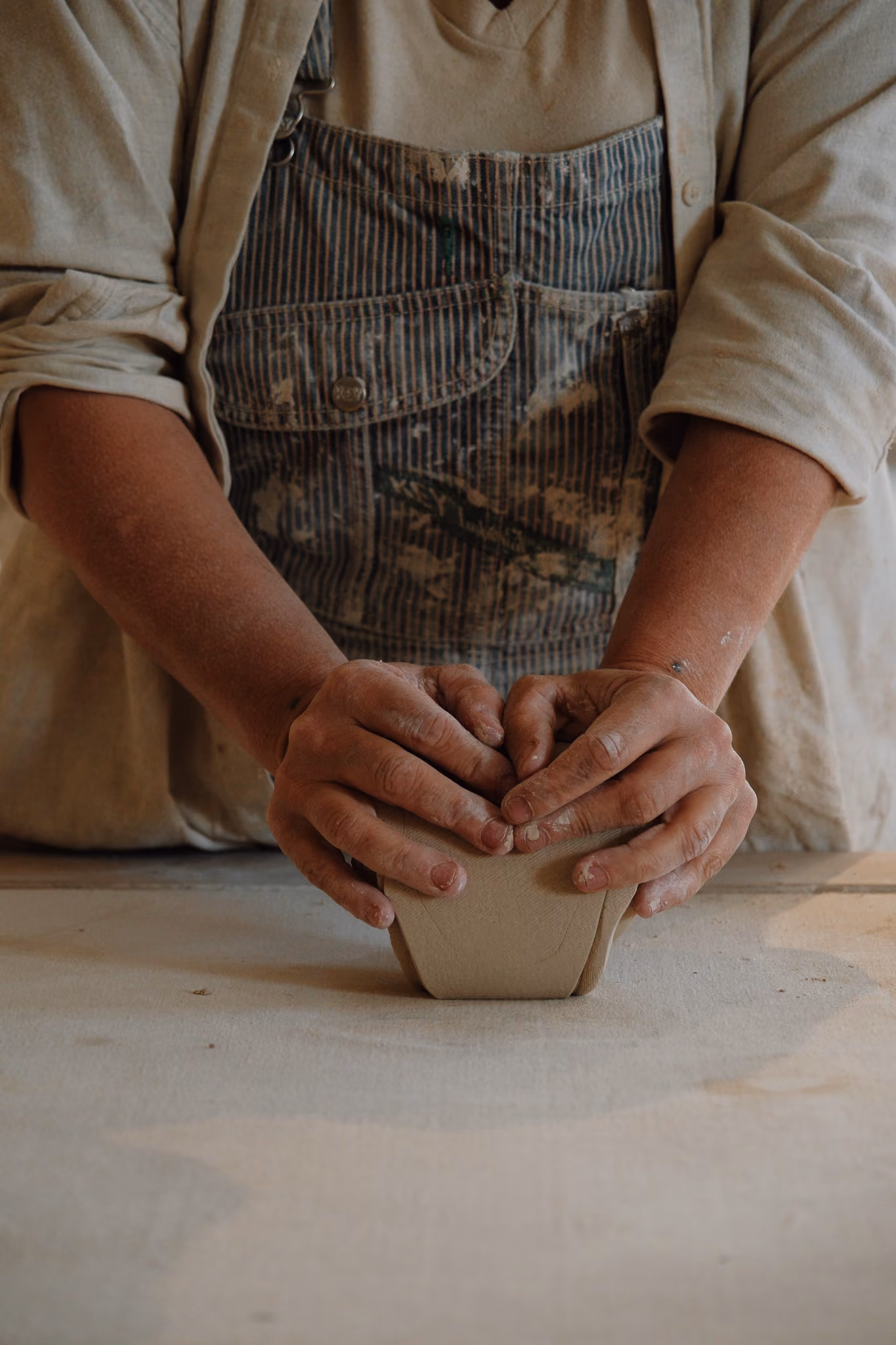 Hands of a person wearing paint-stained overalls and a beige shirt shaping a piece of pottery or clay on a flat surface.