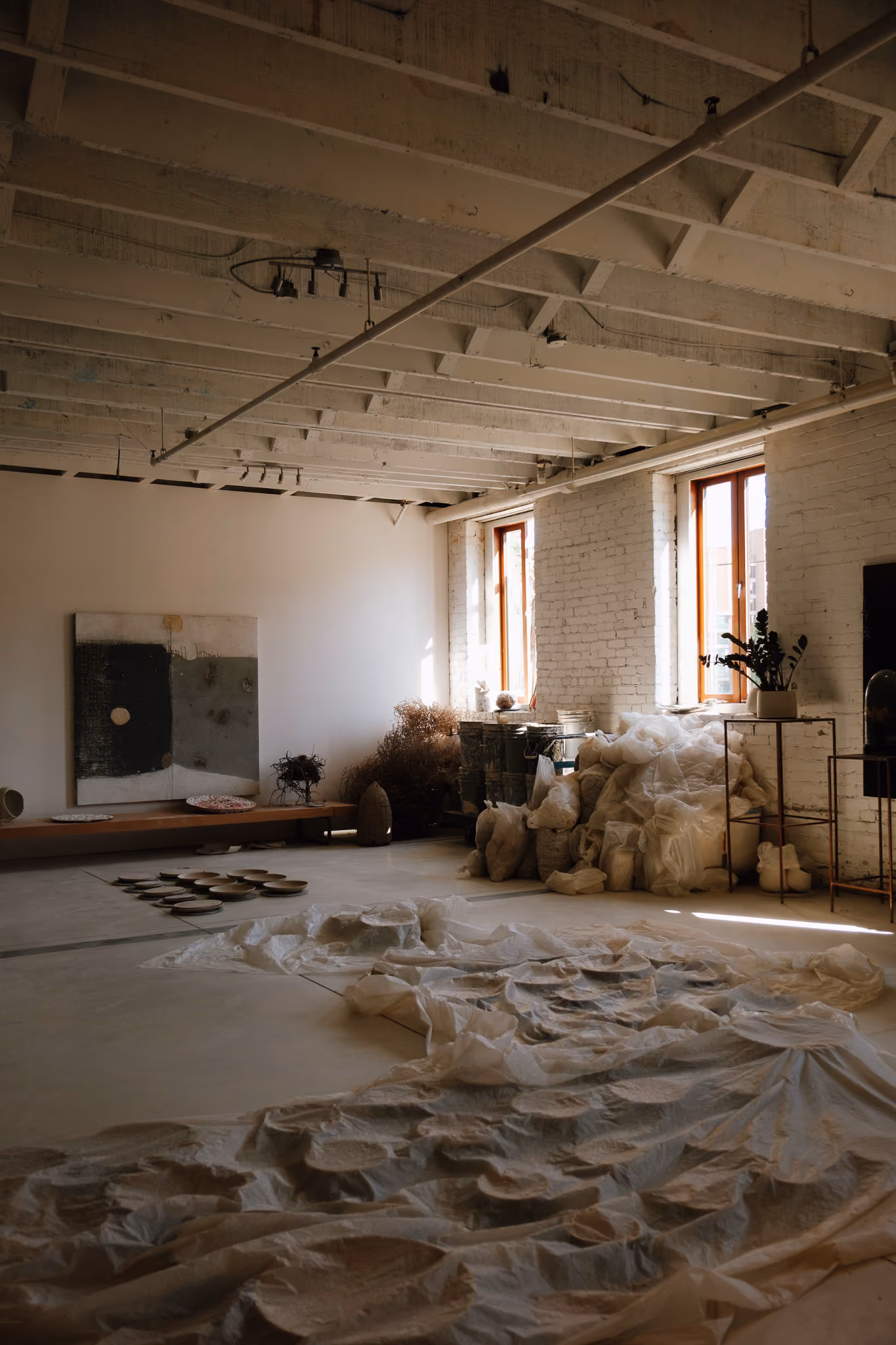 Sunlit art studio with white brick walls and exposed ceiling beams, featuring ceramic plates arranged on the floor covered by translucent white fabric, a low wooden bench with pottery, dried plants, and two large windows letting in natural light.