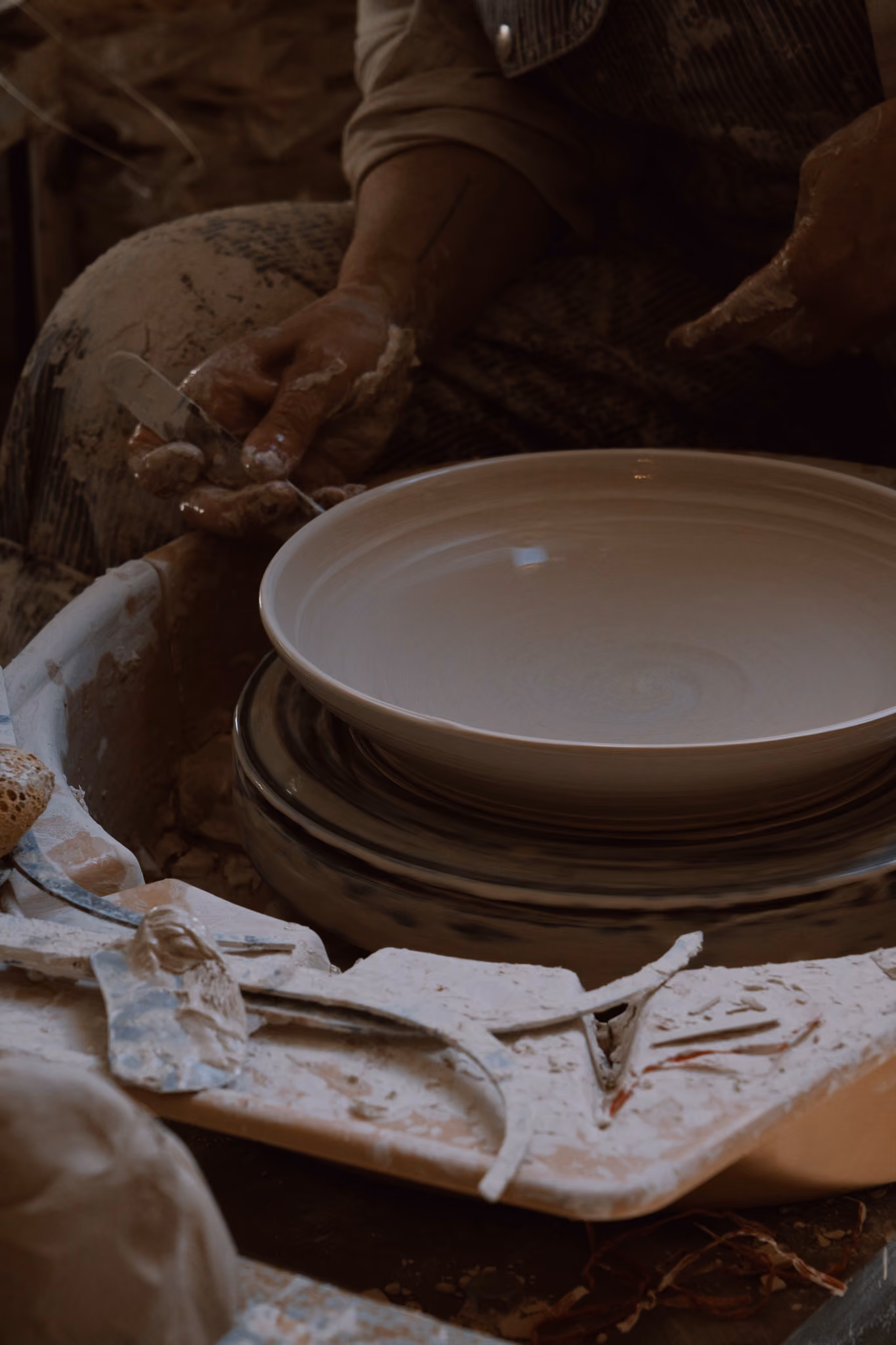 Close-up of a person shaping a ceramic plate on a potter's wheel with clay-covered hands and pottery tools nearby.