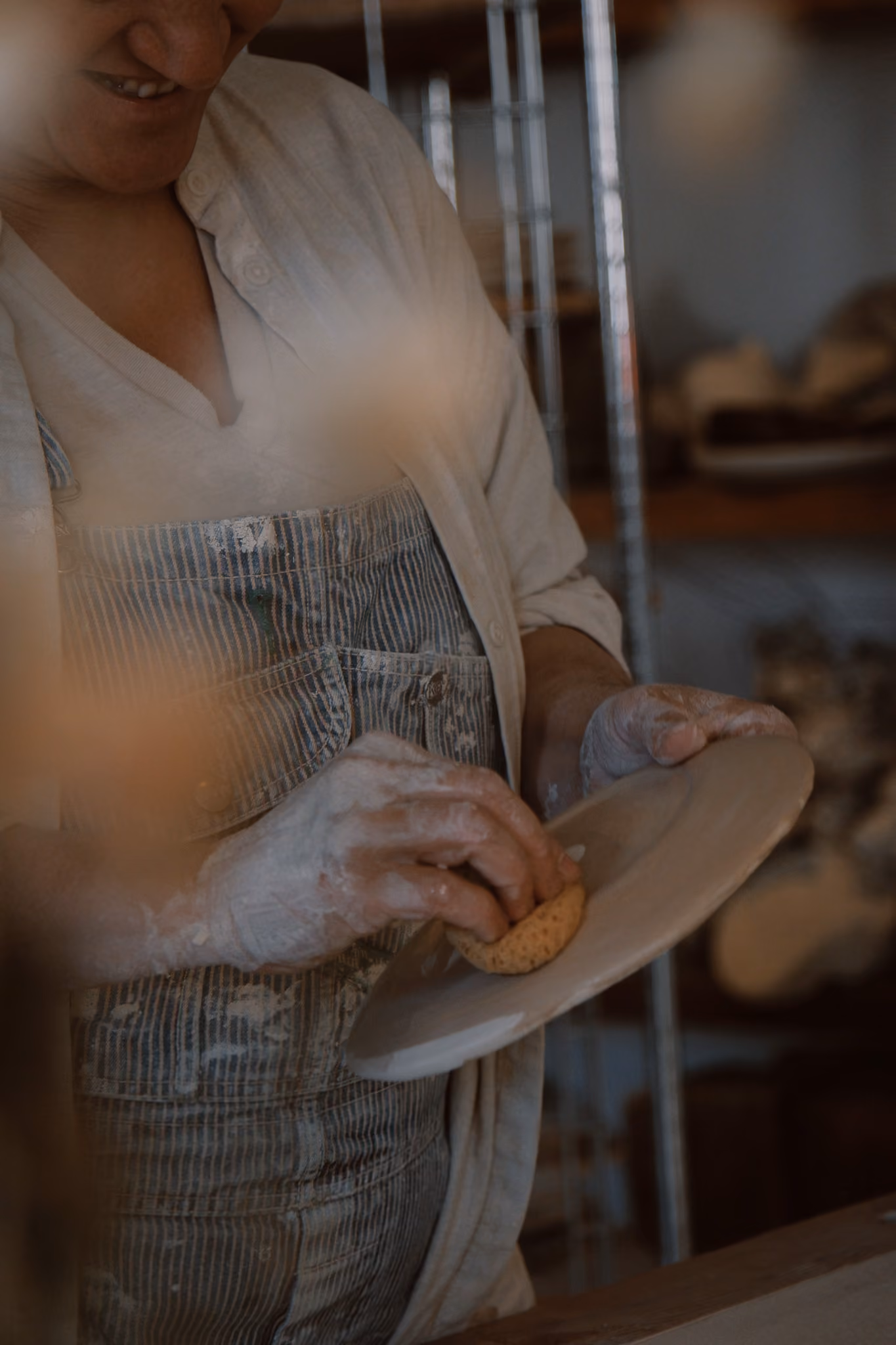 Person wearing striped overalls and a light cardigan, cleaning a clay plate with a sponge in a pottery studio.