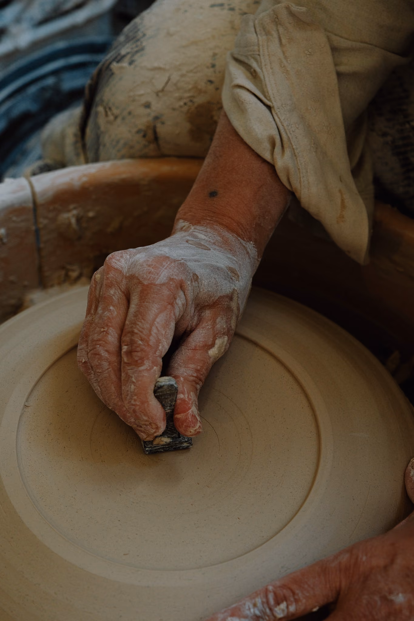 Close-up of a person's hand covered in clay using a small tool to shape the surface of a round pottery piece on a potter's wheel.
