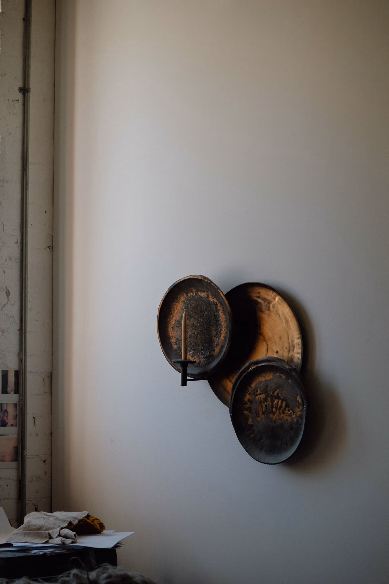 Three rustic, round metal trays mounted on a plain wall, with the left tray holding an unlit beige candle in a simple holder, casting shadows behind them.