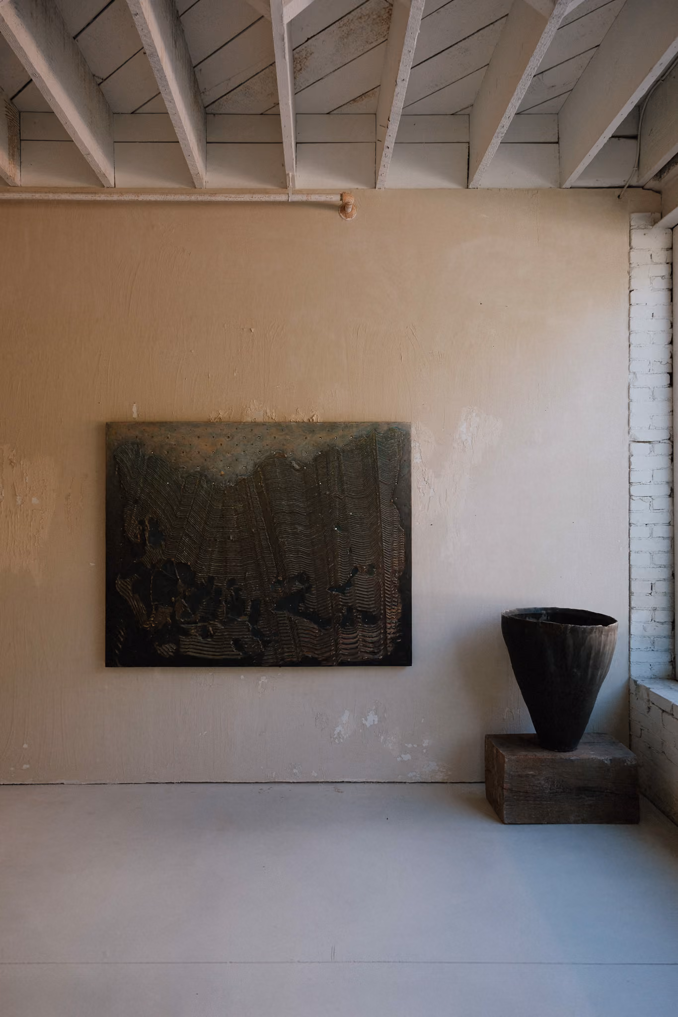Minimalist interior with a textured dark abstract painting on a beige wall and a large dark vase on a wooden block near a window.